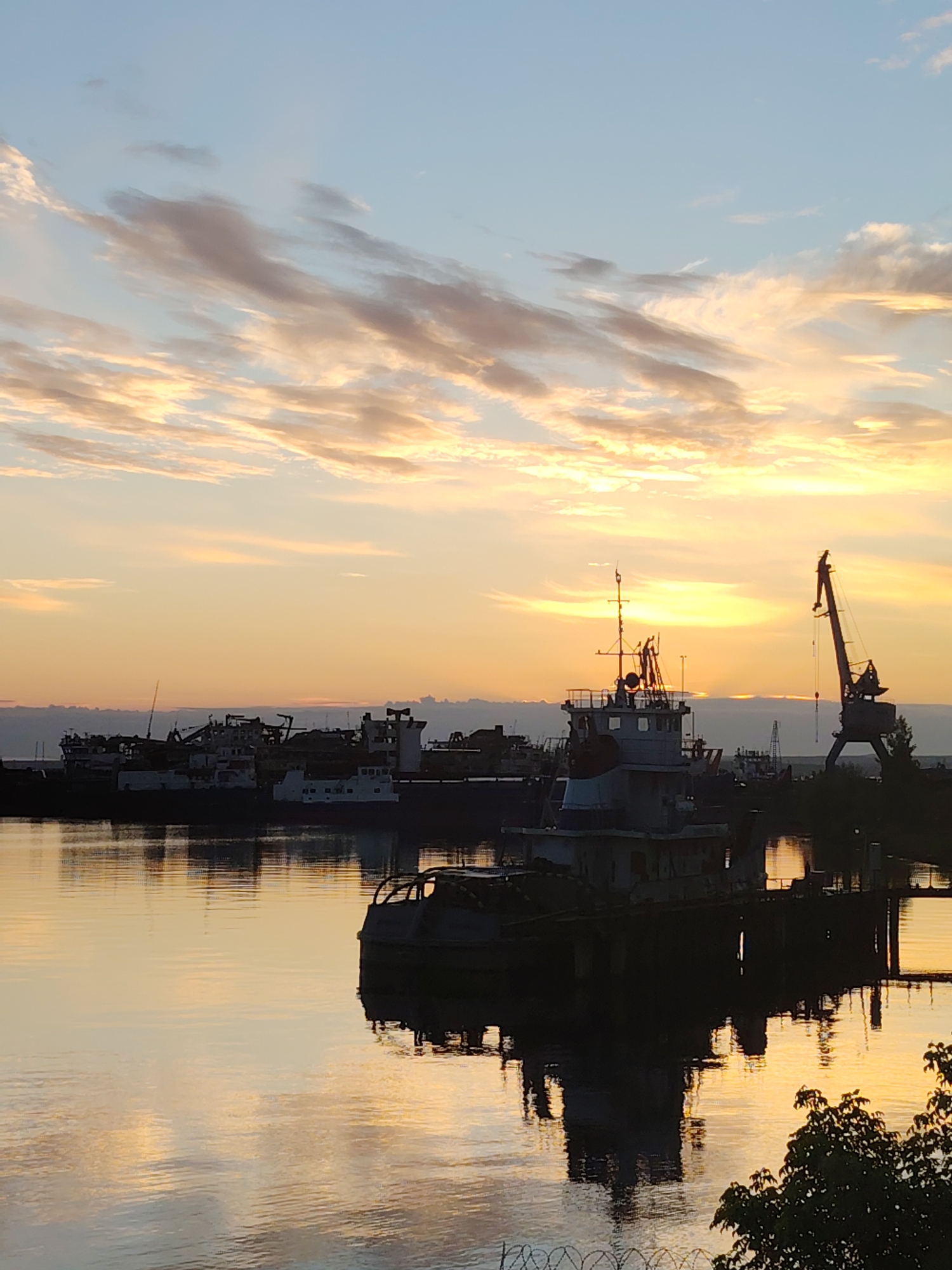 The image depicts a serene harbor scene at sunset in Chaikovsky, Russia. The sky is a canvas of soft blues and oranges, streaked with elongated clouds illuminated by the setting sun. Silhouetted against this vibrant sky are several docked vessels, their forms softened by the twilight. Prominent among them is a tugboat with intricate rigging, its reflection shimmering on the calm water. To the right, a towering industrial crane stands as a stark sentinel, also casting its silhouette onto the water's surface. The harbor appears to be a place of rest for these ships, awaiting their next journey. The overall atmosphere is one of tranquility and stillness, characteristic of the end of a day by the water.