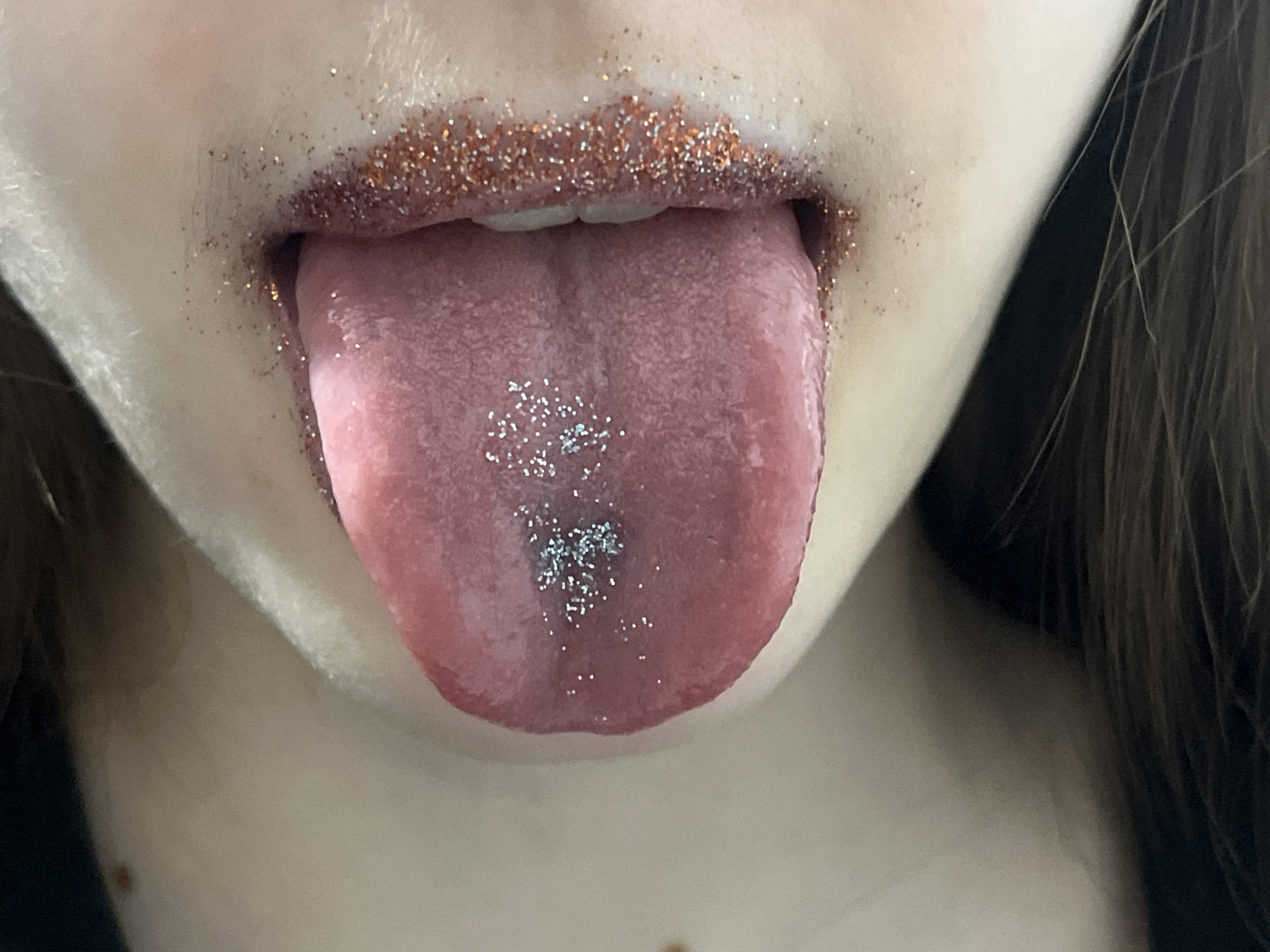 A close-up photograph shows a person's open mouth with their tongue extended. The lips are covered in reddish-orange glitter. There is silver glitter concentrated in the center of the tongue. The individual's skin is fair. The background is indistinct, revealing only partial views of the person's neck and hair. There is no indication of the image being located in Saint Petersburg, Russia.