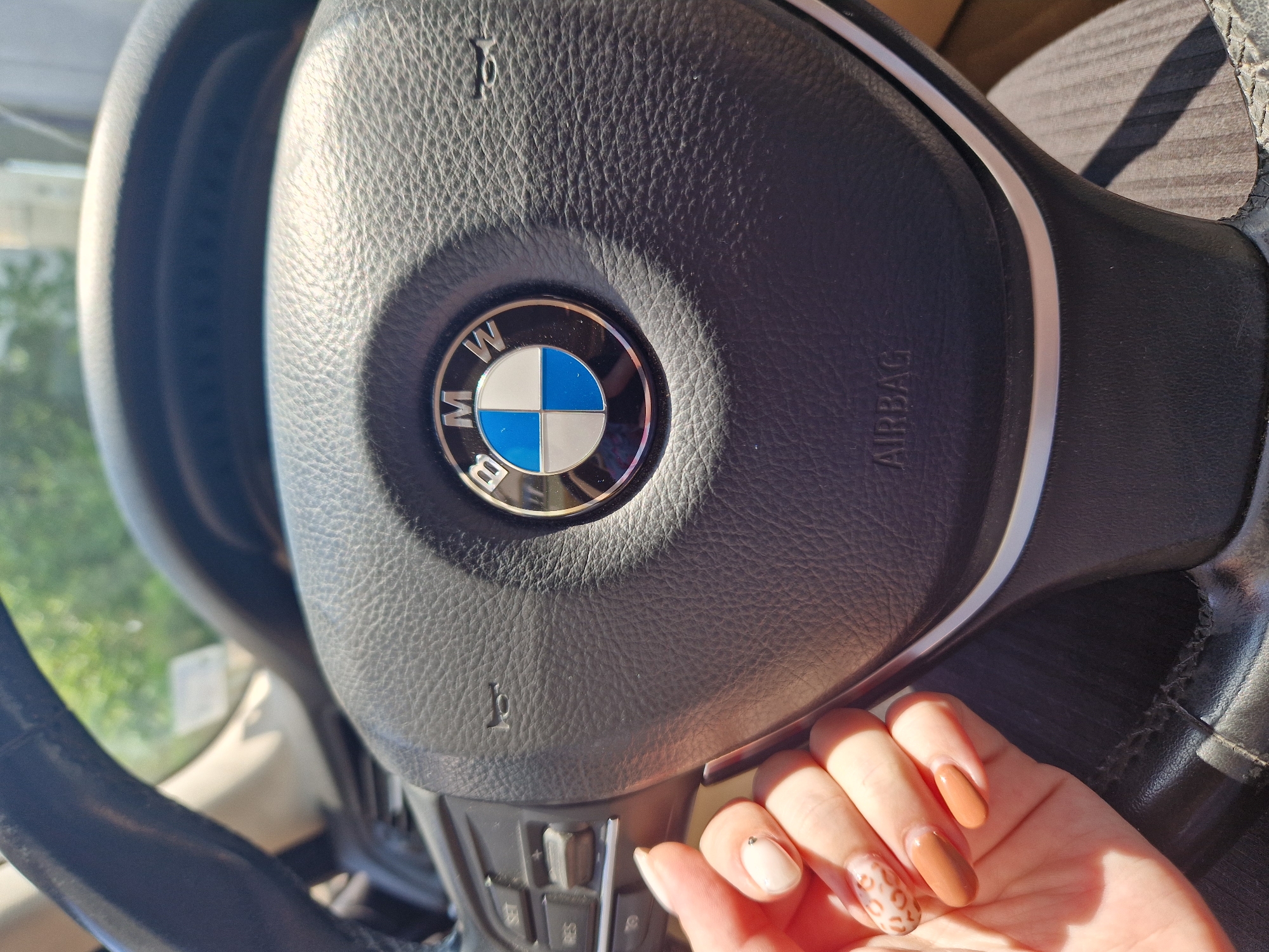 A close-up, high-angle shot captures a person's right hand resting on the steering wheel of a car, likely a BMW, during bright daylight. The steering wheel, made of black textured leather, prominently features the circular BMW logo at its center, displaying the characteristic blue and white quadrants with "BMW" text above and below. To the right of the logo, the word "AIRBAG" is embossed into the leather.
The hand, with manicured nails, is positioned on the lower left side of the steering wheel. The nails display a varied design: the thumb is painted a light, creamy white with a small silver stud near the cuticle; the index and middle fingers are a solid caramel or light brown shade; and the ring finger features a distinctive leopard print pattern in similar brown tones. The pinky finger appears to be a solid light brown.
Below the hand, part of the steering wheel controls are visible, including buttons for "SET" and "RES," suggesting cruise control functions, along with what appear to be volume or media controls. Bright sunlight casts distinct shadows on the steering wheel and the person's hand, indicating a clear, sunny day. Through the car window in the background, out-of-focus green foliage is visible, implying an outdoor setting. The interior of the car, including part of a seat, is visible in the background in soft focus.
