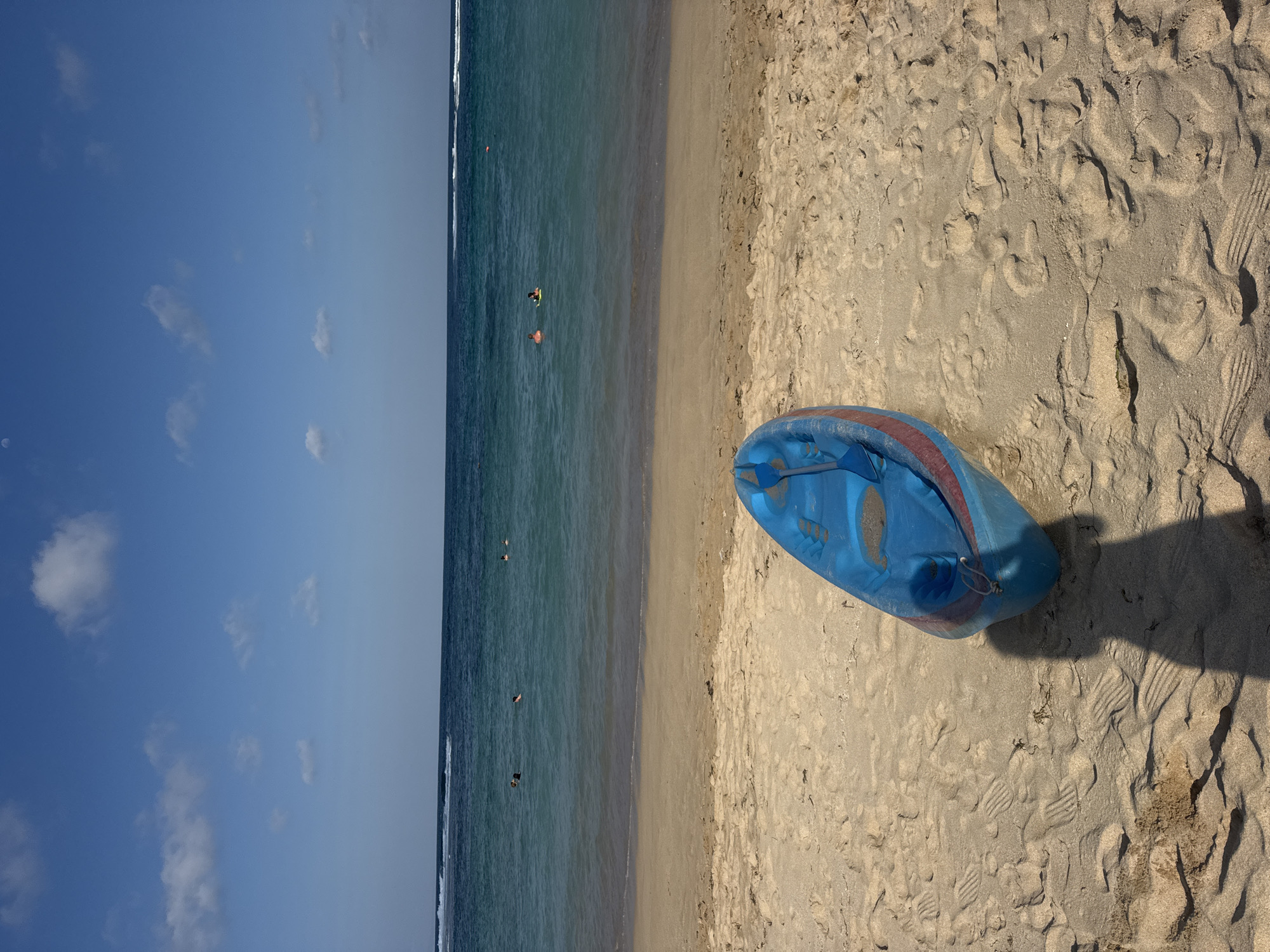 A blue and red kayak rests on a sandy beach, with its front facing towards the viewer. Footprints are visible in the sand surrounding the kayak. The ocean is in the background, with gentle waves breaking on the shore. Several people are visible in the water, some swimming and others on floatation devices. The sky is a clear blue with a few scattered white clouds. The location is identified as Kuta Selatan, Indonesia.