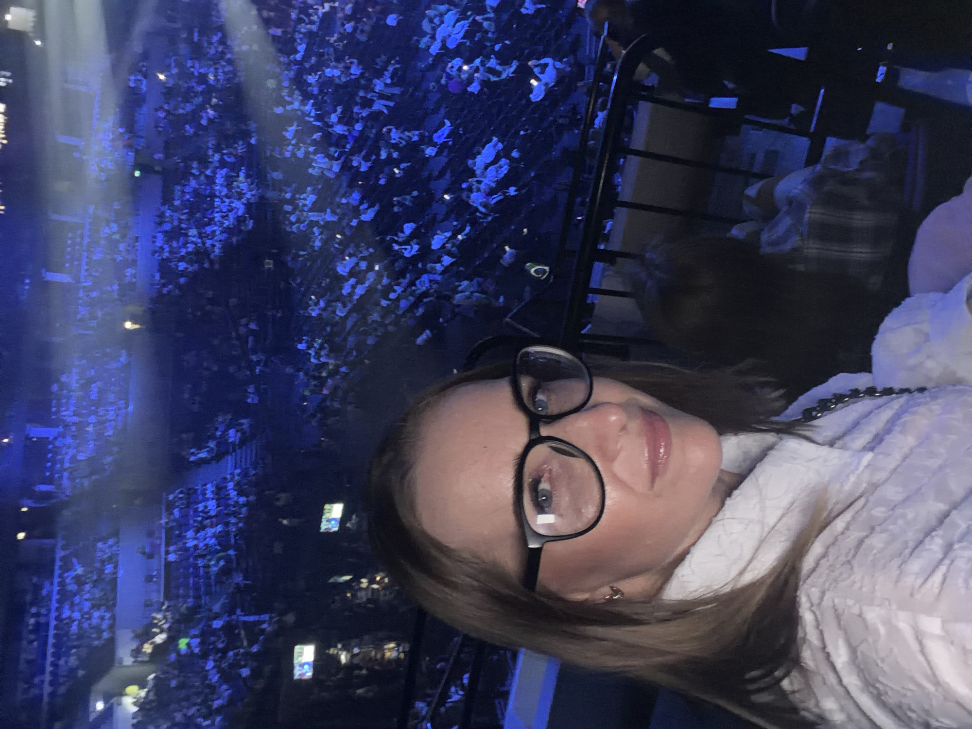 A woman wearing glasses and a white top takes a selfie at a concert in Khimki, Russia. The background features a large crowd of people under blue lighting. A metal railing is visible in the foreground.