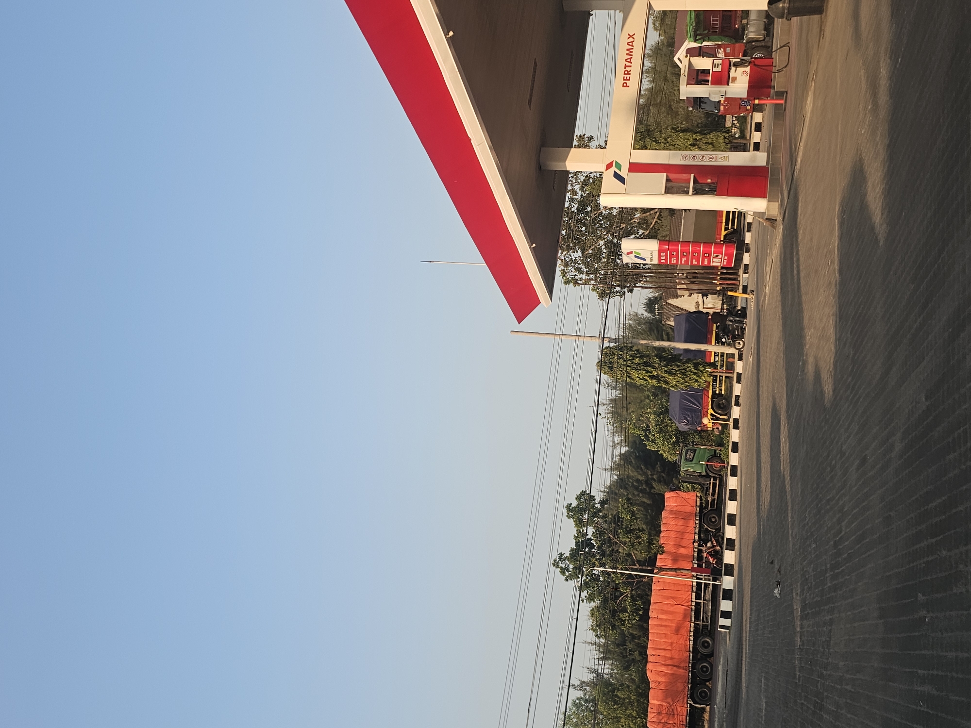 In Jenu, Indonesia, a Pertamina gas station is visible under a clear blue sky. The station features a red and white canopy supported by columns, with "PERTAMAX" signage on the right side. A gas pump with a hose is present. In the foreground, a paved area with a patterned texture is visible. To the left, several trucks are parked. One truck is covered with an orange tarp, and another has a blue tarp. A person is seen riding a motorcycle near the trucks. Overhead, power lines run horizontally across the scene. Trees and other vegetation are present in the background.