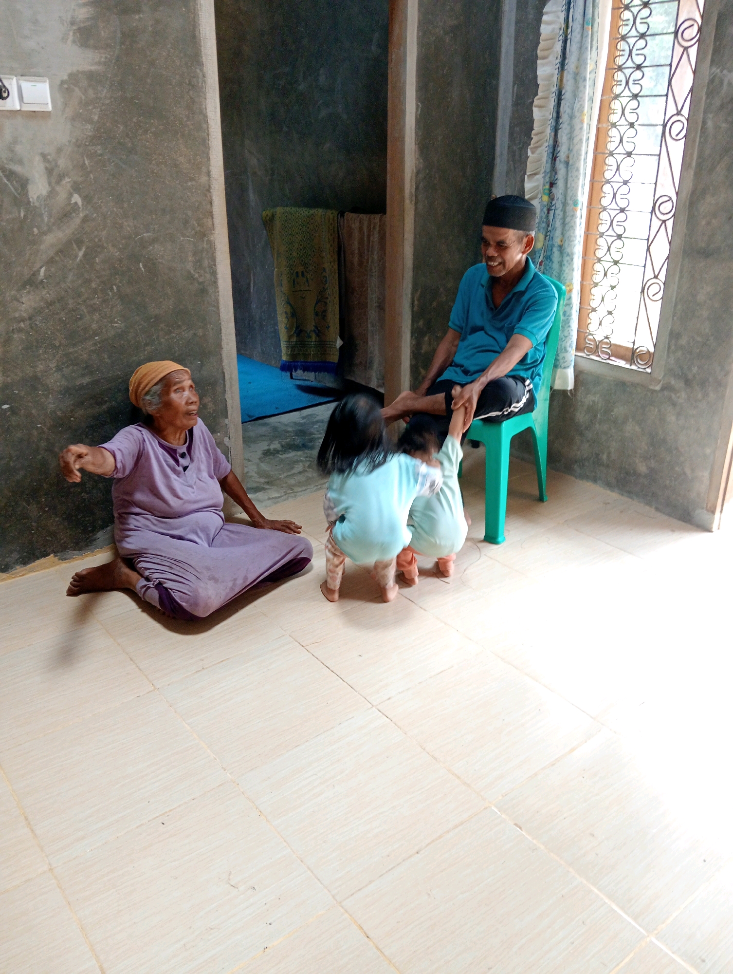 In a dimly lit interior room, possibly a home in Campago Barat, Indonesia, an elderly woman in a purple dress and headscarf sits on the floor, facing left with her left arm extended. A man wearing a blue shirt and a black peci (cap) sits on a green plastic chair to the right, smiling. Two small children, one in a light blue outfit and the other in a similar light-colored outfit, are interacting with the man. One child is holding his hand, while the other appears to be kneeling. The room has textured concrete walls, a tiled floor, and a window with decorative metalwork and curtains on the right. A patterned cloth hangs in the background on the left. The scene suggests a moment of family interaction.