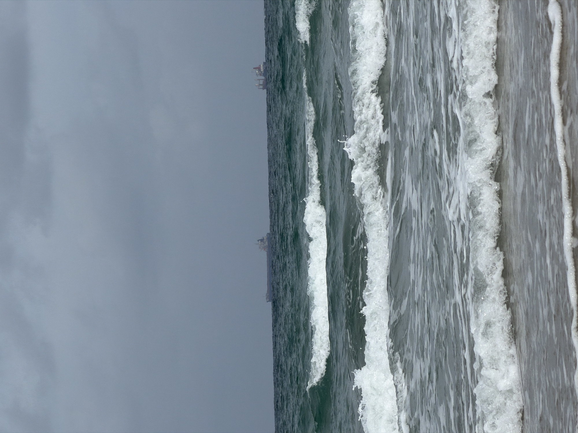 The image depicts a view of the Mediterranean Sea in Kiryat Yam, Israel, under an overcast sky. Waves with white foamy crests are prominently displayed, moving from the ocean towards the shore. In the distance, a ship and offshore platforms are visible on the horizon. No people are visible.