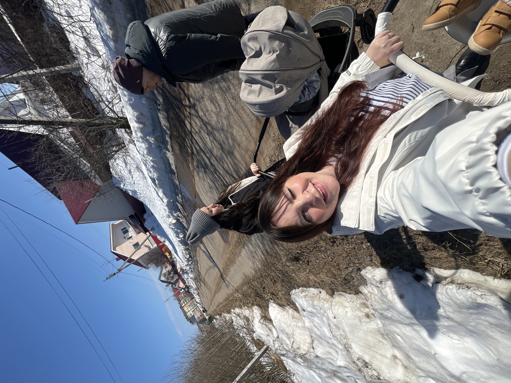 The image displays three individuals, primarily women, walking along a dirt road in Noviy, Russia, under a clear blue sky. The season appears to be late winter or early spring, characterized by patches of melting snow and bare vegetation along the roadside.
In the immediate foreground, a woman with long, dark brown hair and a white jacket smiles directly at the camera, holding the handle of a grey baby stroller, indicating a selfie perspective. She is wearing a striped shirt beneath her jacket. To her right and slightly behind, a second woman, wearing a grey knitted beanie and a black jacket, is also present near a stroller, looking forward and to the right. A third individual, clad in a dark beanie and a dark puffy jacket, walks further to the right in the background, also facing forward.
The road itself is unpaved, showing wet, muddy sections and tire tracks. Large drifts of dirty, melting snow line the edges of the road, interspersed with brown, sparse vegetation. In the background, several residential structures are visible, including a light pink house and another with white siding, both featuring red roofs. Power lines stretch across the upper part of the image, supported by a utility pole on the left. Bright sunlight illuminates the scene, casting discernible shadows.