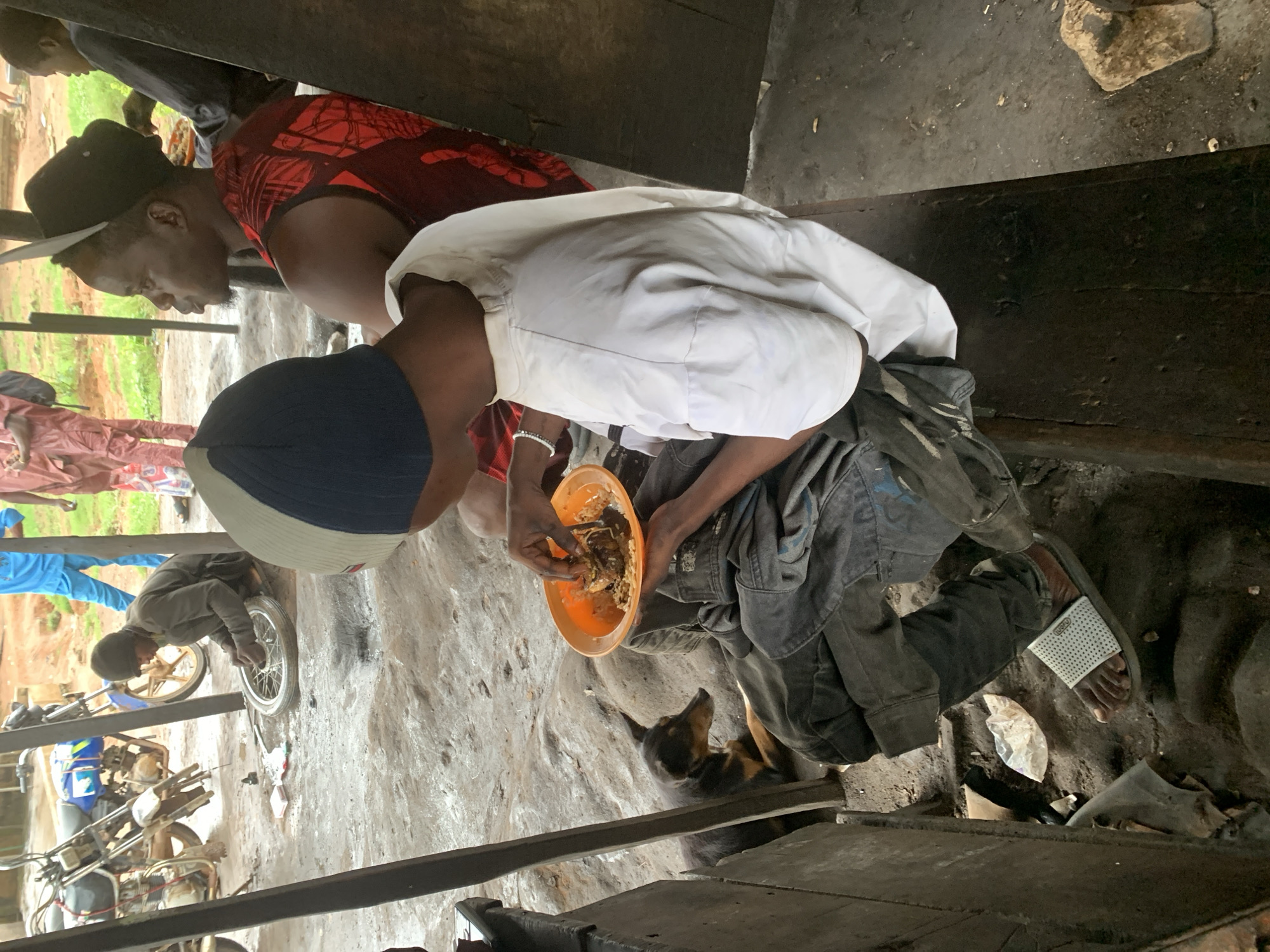 In Shagamu, Nigeria, within an open-air workshop area with a dirt floor, a young man wearing a white t-shirt and dark trousers, with a blue and grey cap, is seated on a wooden bench. He is in the process of eating from an orange bowl held in his left hand, using his right hand to pick up food. A small dog with dark fur lies on the ground to his left. To his right, another young man in a red sleeveless shirt is seated. In the background, a motorcycle is visible, and other individuals are present, some wearing traditional Nigerian attire. The environment appears to be a functional workshop, possibly a mechanic's shop, indicated by tools and parts scattered on the ground and a bicycle wheel being worked on by another person not fully in frame.