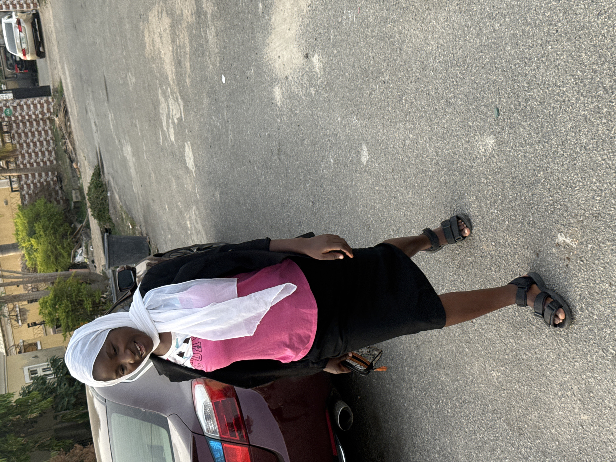 A young person, likely female, stands smiling broadly on a paved road next to a parked car. She is dressed in a white headscarf, a bright pink t-shirt with stylized text, a black long-sleeved top or jacket, and black open-toed sandals. In her right hand, she holds a small, patterned wallet or phone holder.
The setting is outdoors in Abuja, Nigeria, on what appears to be a street or driveway. A dark-colored car, possibly maroon, is parked behind her, with its rear lights visible. The road surface is asphalt, showing some signs of wear and lighter patches. In the background, there are various buildings with light-colored facades and dark roofs, interspersed with green trees and bushes. A patterned fence, possibly white and brown tiles, is visible in the middle ground, and another white car is parked further down the road.
The lighting suggests a bright, clear day, possibly late afternoon or early evening, casting soft shadows. The person's cheerful expression indicates a positive emotion while posing for the photograph in this casual outdoor environment.