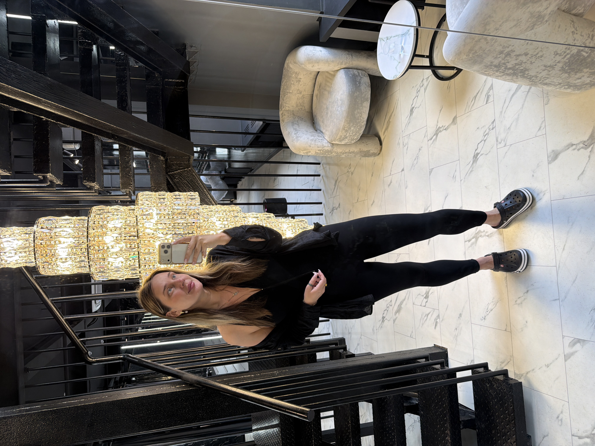 A woman stands on a marble-tiled floor, taking a mirror selfie. She is wearing black leggings, black Crocs, and a black top with a black cardigan draped over her shoulders. She has long brown hair and is wearing earrings and a necklace. In the background, there is a black metal staircase with diamond plate steps, a tall, multi-tiered crystal chandelier, and a light gray armchair next to a small circular table with a glass of water. The location is stated to be in the village of Ukromne, Simferopol Raion, Crimea.