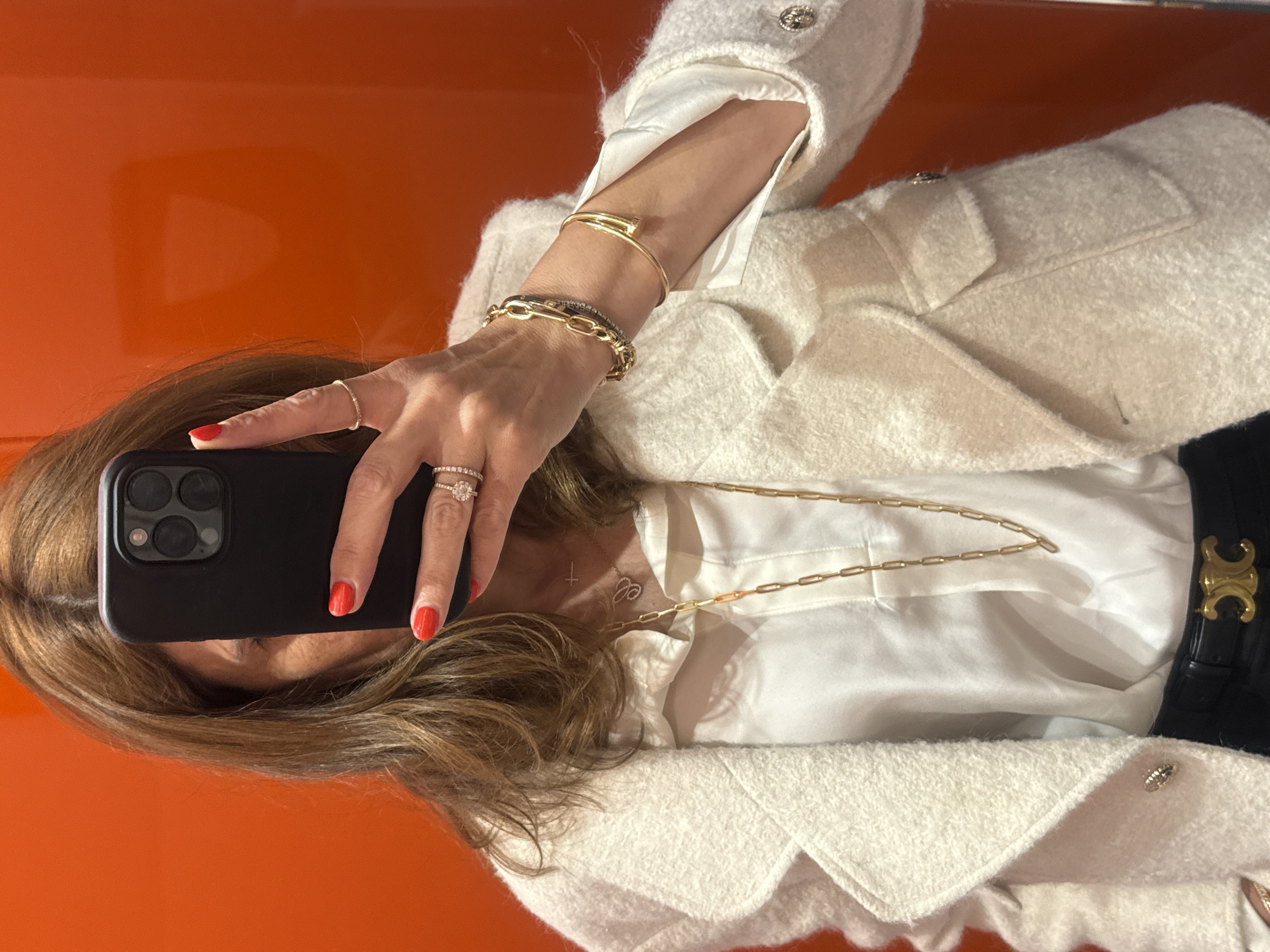 A woman takes a mirrored selfie, holding a black smartphone with her right hand and obscuring her face. Her nails are painted red. She wears multiple rings on her right hand and several bracelets on her left wrist, including gold bangles and a chain bracelet. She's wearing a white collared shirt under a textured white jacket with decorative buttons. A gold chain necklace hangs over the shirt. A black belt with a gold-toned buckle is visible at her waist. The background behind the mirror is a solid, bright orange. There is no visible evidence to suggest the location is Lisbon, Portugal.