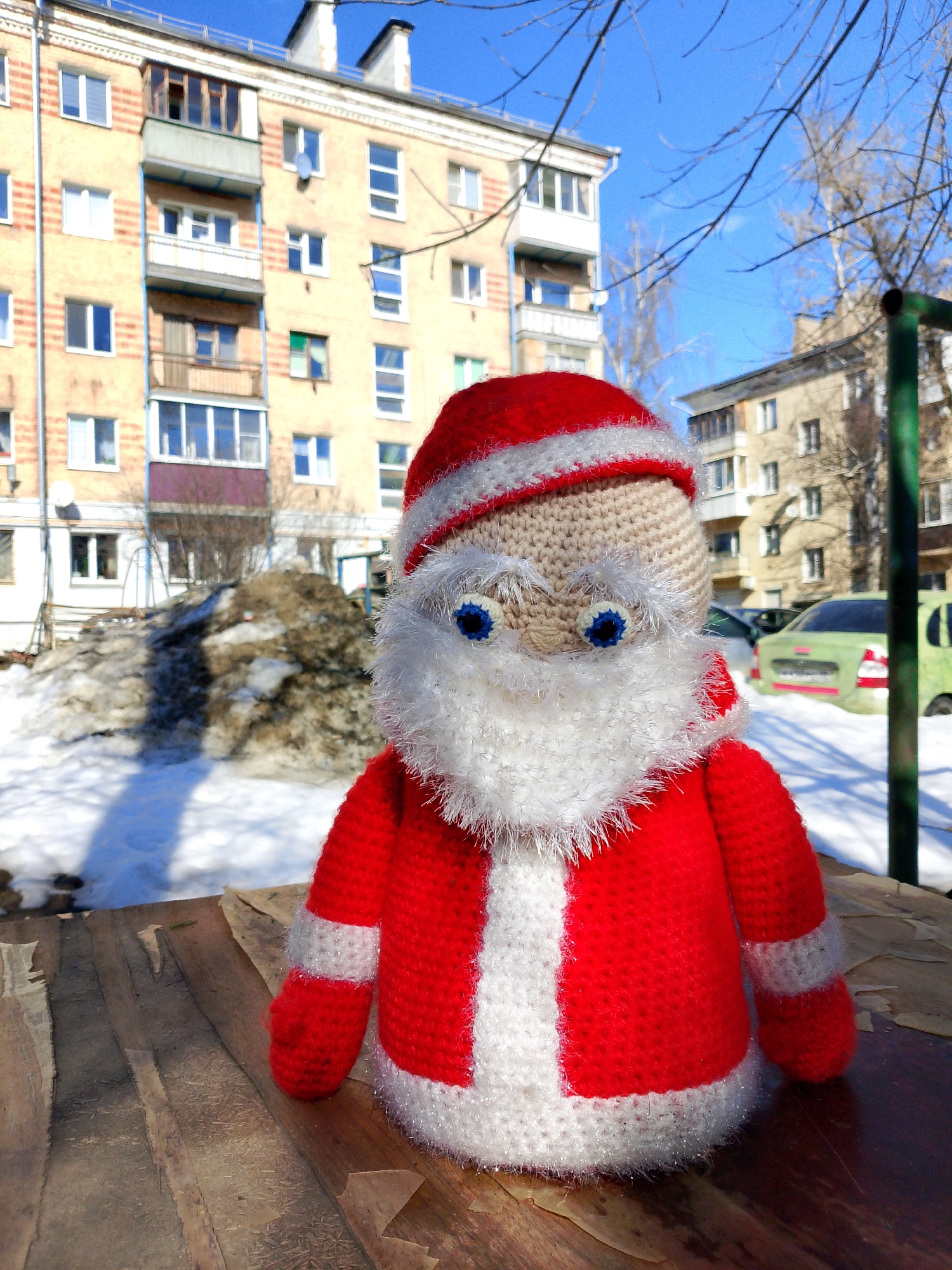 The image displays a crocheted Santa Claus doll positioned in the foreground, resting on a worn, light-brown wooden surface. The doll features a beige head, blue yarn eyes, and a white fuzzy yarn beard, eyebrows, and trim on its red suit and hat.
The setting is an urban environment in Izhevsk, Russia, observed during winter or early spring conditions. The ground in the midground is largely covered with snow, including a prominent dirty snow pile situated to the left of the doll. A clear blue sky is visible overhead.
In the background, multiple multi-story residential buildings are visible. The primary building on the left exhibits a light yellow facade with vertical red stripes, numerous windows, and several balconies, some enclosed. Bare tree branches extend into the upper right portion of the frame. To the right, a second building is partially obscured by trees and a green metal structure. Several parked cars, including a light green sedan, are discernible on a snow-covered street or parking area near the background buildings.