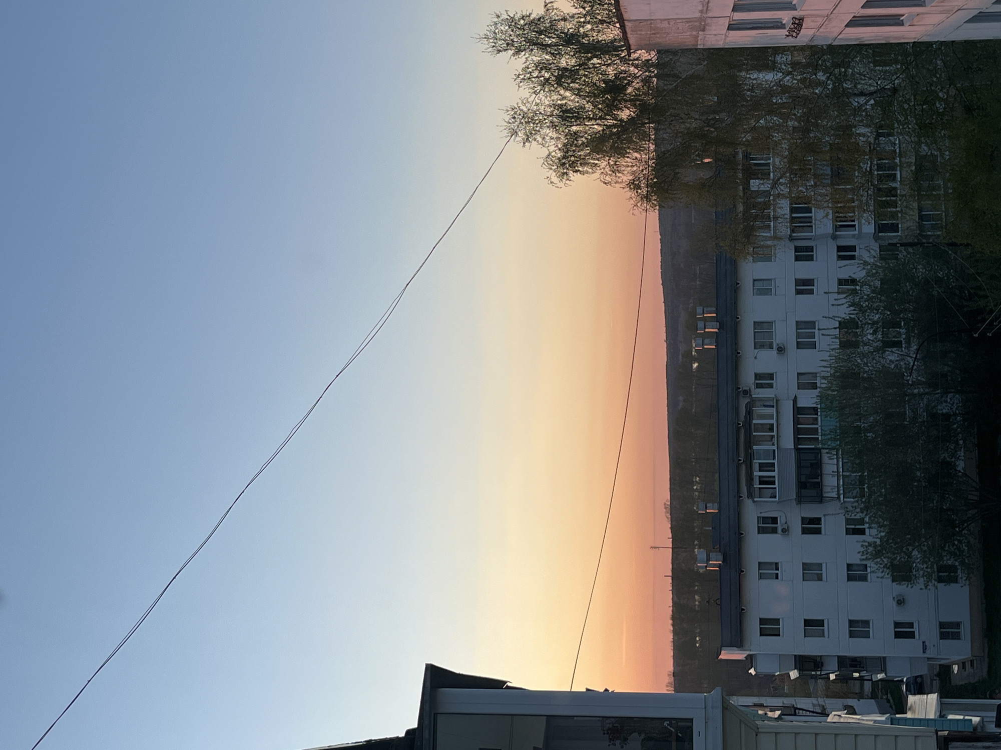 The image captures a cityscape during either sunrise or sunset in the city of Chaikovsky, Russia. The sky transitions from a light blue to a warm orange hue near the horizon. A multi-story white apartment building occupies the right side of the frame, with a number of windows visible. Tree branches are visible both near the building and partially obscuring the horizon. In the foreground, there is a building that is obscured by a roof in the center. An electrical wire stretches diagonally across the sky. The shot is from a slightly elevated viewpoint.