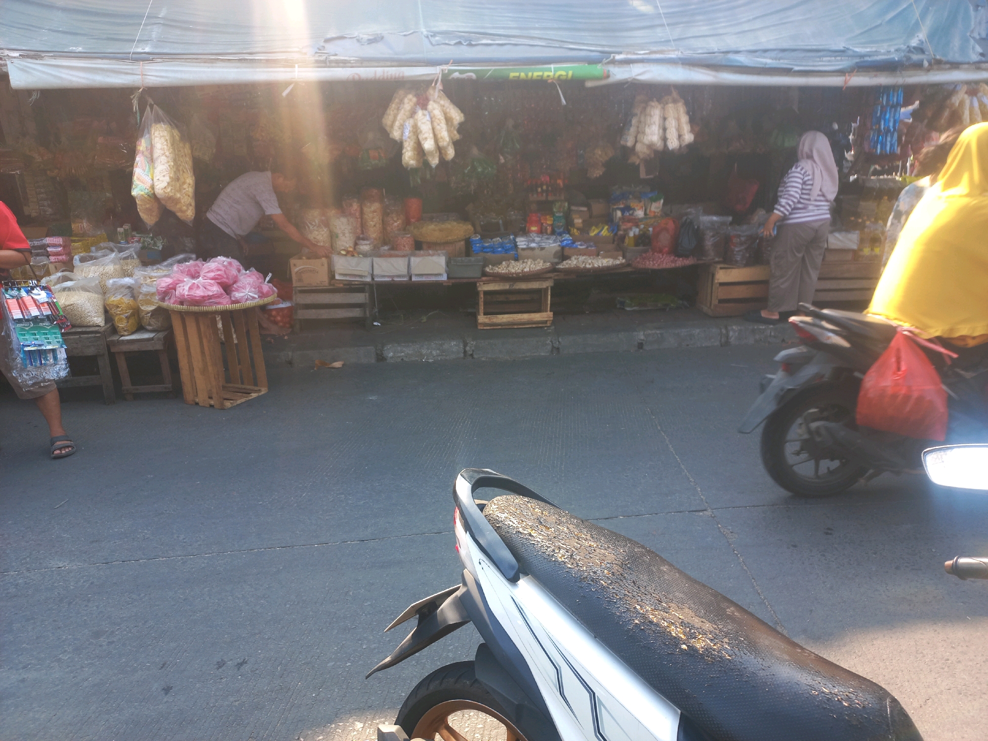 A street-level, eye-level, medium shot captures a bustling outdoor market scene, bathed in bright sunlight. In the foreground, the seat of a silver and white motorcycle is covered in dirt and debris, with a portion of its black handlebar visible. To the right, a motorcycle with a red bag is parked. The middle ground features a row of market stalls with awnings providing shade, displaying various goods such as packaged snacks, bags of food, and bottled items. Two individuals are present: a man in a gray t-shirt is seen behind a stall, appearing to arrange items, and a woman in a striped long-sleeved shirt and hijab is standing at another stall. The ground is paved with concrete, marked by a distinct crack running across its surface. The overall impression is that of a vibrant local market in Depok, Indonesia, during daylight hours.