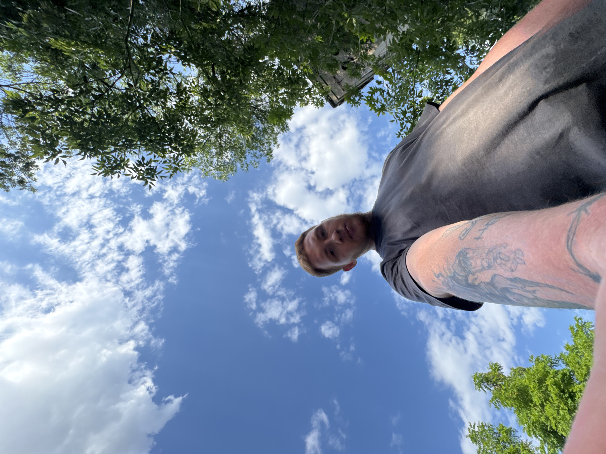 A low-angle, wide shot captures a person from the perspective of looking up from the ground. The person, with reddish-brown hair, is wearing a dark t-shirt and is positioned in the lower right quadrant of the frame. Their right arm is extended, revealing a tattoo on their bicep. Above, a bright blue sky is dotted with white, fluffy clouds. Green foliage from trees frames the top and right sides of the image. A weathered, concrete structure, possibly part of a building, is visible through the leaves in the upper right. The lighting suggests it is daytime, with the sun filtering through the leaves. The scene is outdoors, under a clear sky, with vegetation and a man-made structure present.