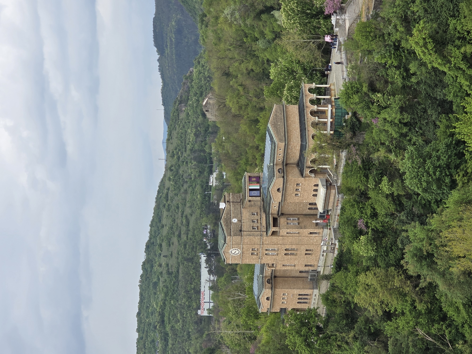 The image presents a panoramic view of a large, multi-structured building complex situated at the base of a heavily forested mountain range under an overcast sky. The buildings are constructed from light brown or beige stone/brick, featuring dark-colored roofs, numerous windows, and sections with arched colonnades and glass skylights.
The setting is lush with dense green foliage, particularly noticeable on the steep mountainside and surrounding the buildings. The varying shades of green suggest spring or early summer, with some trees displaying pinkish-purple blossoms. A paved pathway or terrace runs alongside the buildings, where several small figures are visible, indicating human presence.
Notable details include at least two prominent clock faces on different towers or sections of the buildings. On the tallest section of the main building, two flags are flying; one is clearly the European Union flag, while the other features red and yellow/gold colors. In the distance, partially obscured by trees, a large white sign with text in Cyrillic is visible, reading "ТЕХНОПОЛИС" (TECHNOPOLIS). The weather appears overcast, with soft, diffused light, suggesting it is daytime. The overall scene depicts a blend of architectural development and natural beauty within a mountainous, verdant landscape.