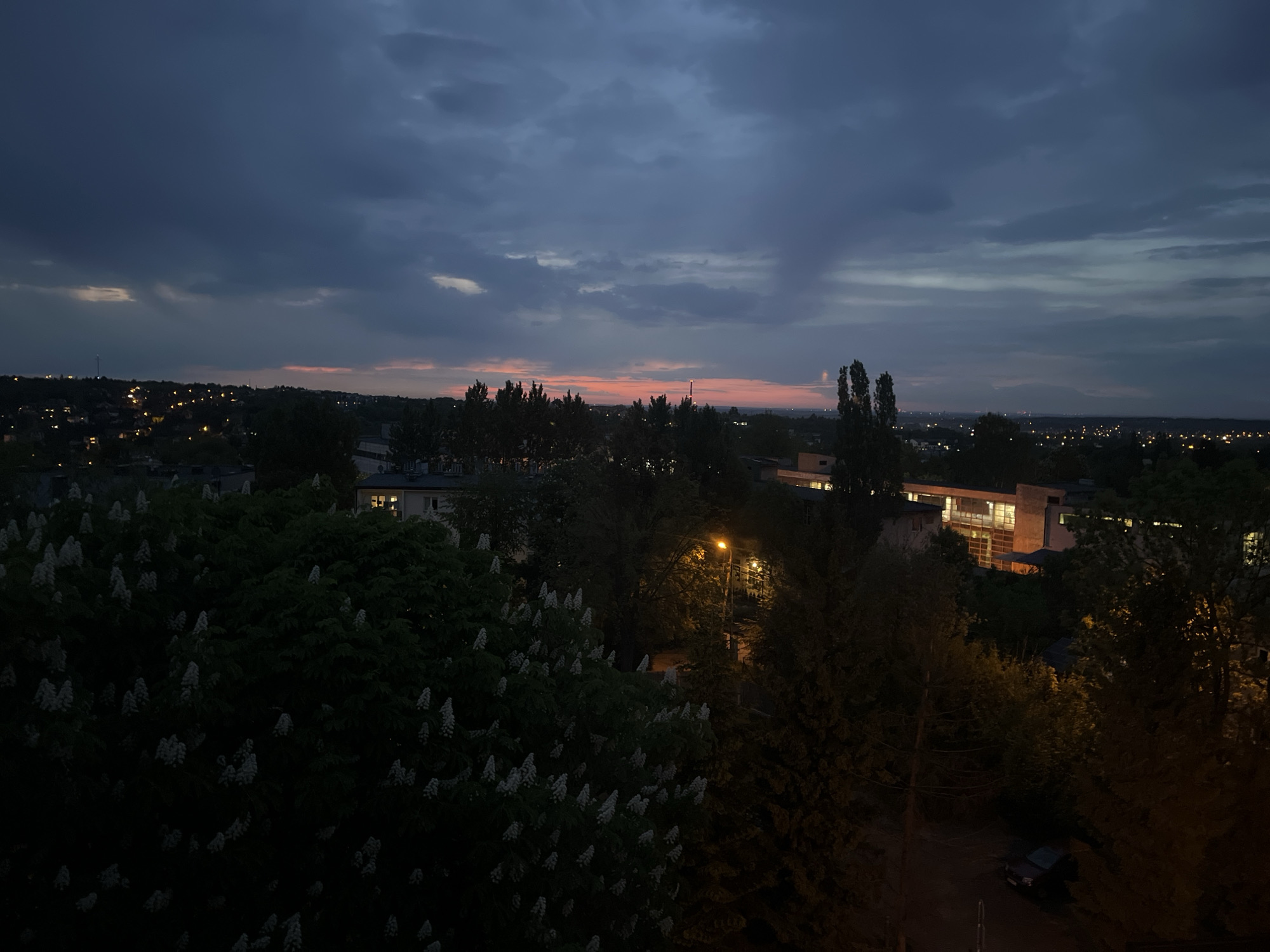 Here is a detailed description of the image:
**Environment:** The scene is an elevated view of an urban area at dusk or twilight. The sky is dominated by dark, heavy clouds, with a faint orange glow along the horizon indicating the setting sun.
**Urban Landscape:** The landscape is populated with buildings of varying sizes and designs, interspersed with trees and vegetation. There are scattered lights visible in the distance, suggesting residential areas and infrastructure.
**Foreground:** The foreground includes dense foliage, possibly a garden or park, with trees and flowering bushes.
**Objects:** There is a street lamp with an orange glow, illuminating some of the surrounding trees. A car is parked below, near some of the greenery.
**General Impression:** The scene conveys a sense of tranquility and a quiet urban atmosphere at the transition between day and night.