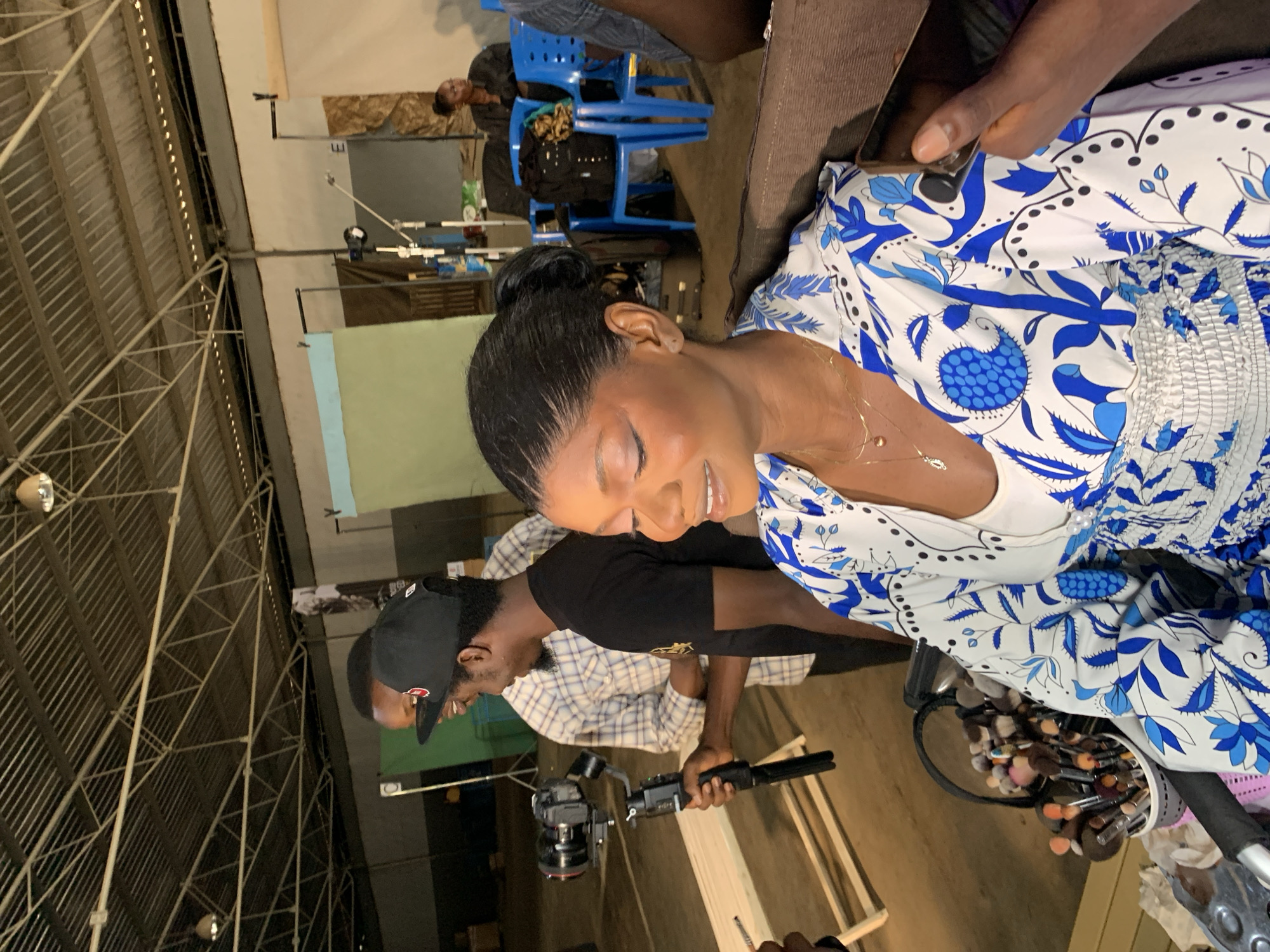 A group of people are gathered in what appears to be a studio or production setting in Barakin Lerawi, Nigeria. In the foreground, a woman with her eyes closed is being attended to by someone whose hand is visible near her face, possibly applying makeup. She is wearing a white dress with a blue floral pattern and a delicate necklace. To her left, a man wearing a black baseball cap and a plaid shirt is holding a professional camera with a stabilizer. Behind them, more people are visible, with some seated on blue chairs in the background. A large industrial-style ceiling structure with exposed beams and lighting is prominent in the upper portion of the image. There is a collection of makeup brushes in a container to the right of the woman. The scene suggests a behind-the-scenes look at a photo or video shoot, with the focus on the woman and the production equipment. The lighting appears to be artificial, and the overall atmosphere seems focused and professional.