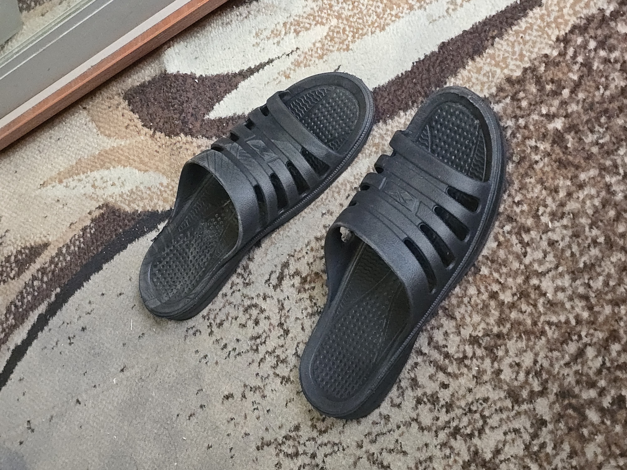 Two black slip-on sandals are placed on a carpet. The sandals are made of a flexible material and have a textured footbed. The carpet has a patterned design with brown, beige, and white stripes. The scene appears to be indoors.