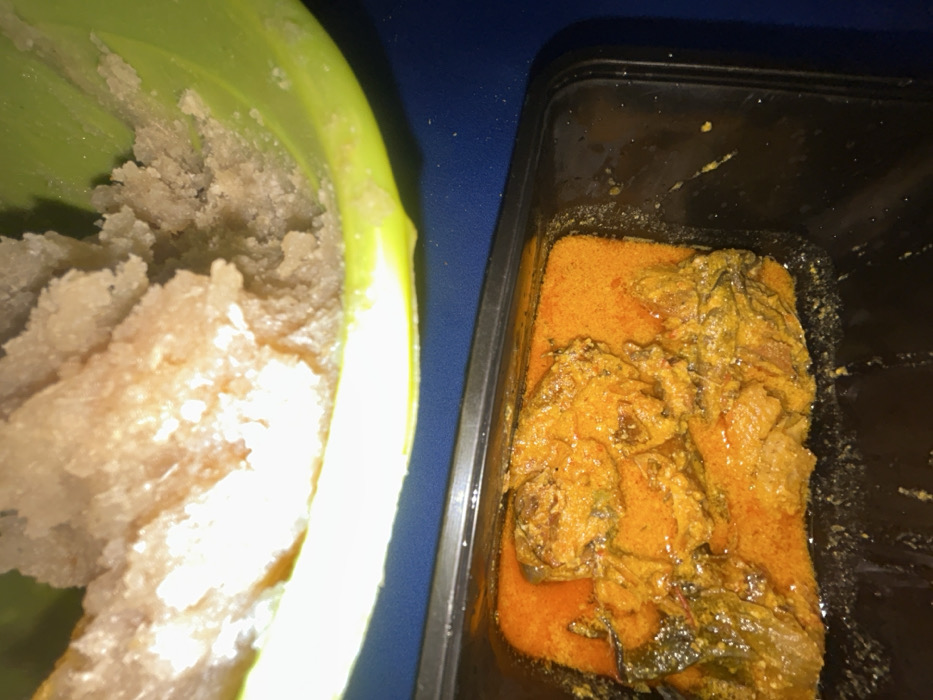 The scene depicts two distinct food preparations arranged on a dark blue surface. In the upper portion of the frame, a light green plastic bowl, partially visible, contains a quantity of light brown, granular, and somewhat lumpy food substance, consistent with a Nigerian 'swallow' staple. Directly below this, a black rectangular plastic container holds an orange-red stew. The stew is thick and contains multiple pieces of protein, likely fish or meat, along with darker, possibly leafy green, elements. No individuals or specific actions are visible. The setting is an indoor environment within the City of Ilishan, Nigeria.