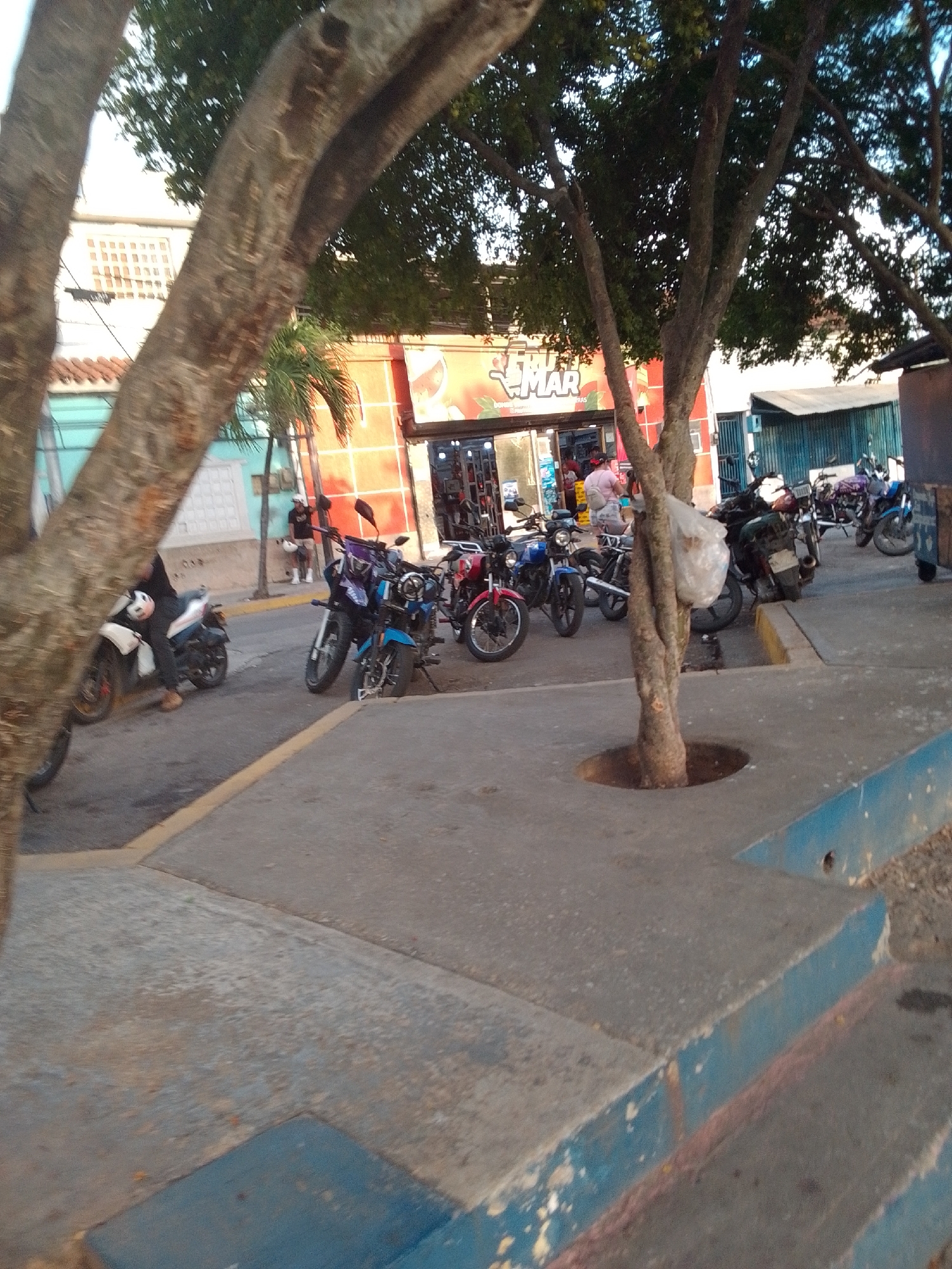 The image depicts a street scene in Juan Griego, Venezuela, featuring a row of parked motorcycles and a store with an awning. In the foreground, several motorcycles are visible, some with riders. The store, identified by a prominent "MAR" sign, appears to be a commercial establishment. The environment suggests a typical urban setting with buildings and a sidewalk. The lighting indicates it is daytime.