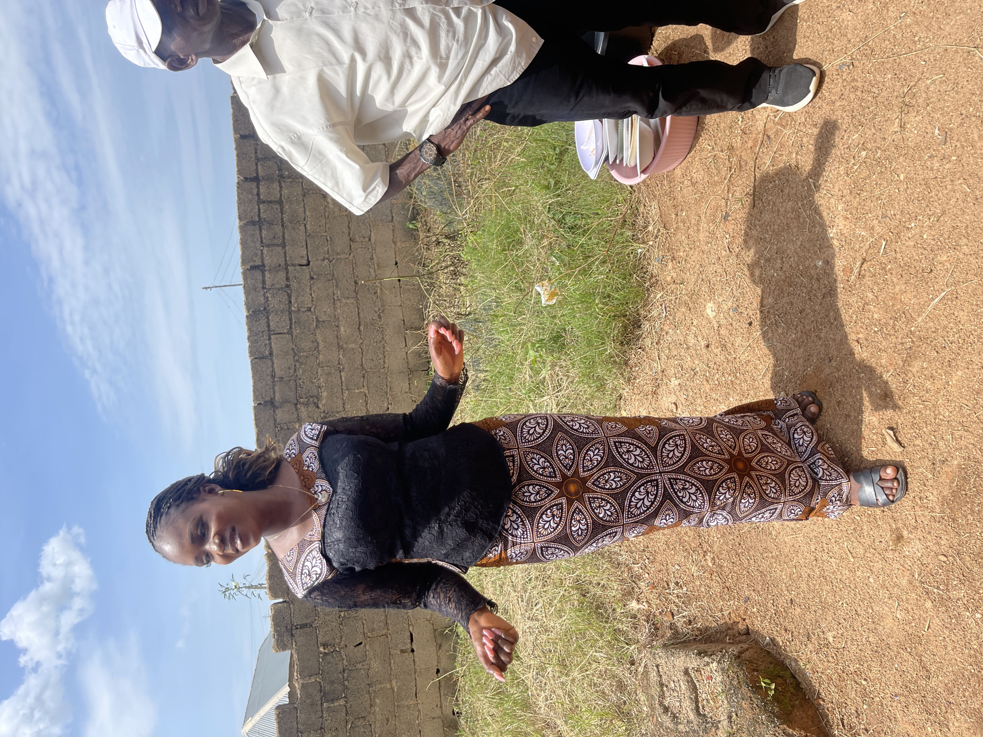 Two individuals, a woman and a man, stand outdoors in what appears to be Rafin Par, Nigeria. The woman, in the foreground, is wearing a long, patterned skirt and a black lace top with long sleeves. She has braided hair and is looking to her left. The man, to the right of the woman and slightly behind her, is wearing a white button-down shirt, black pants, and a white baseball cap. He has his left hand on his hip. In the background, a low brick wall runs horizontally across the scene, with dry grass and some sparse vegetation growing behind it. The ground in the foreground is reddish-brown dirt with scattered dry grass. A pink basin containing several white bowls is visible near the man's feet. The sky is blue with scattered white clouds.