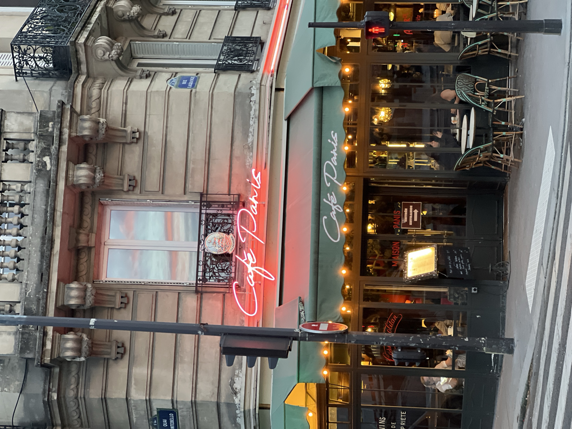 The image presents a vibrant Parisian street scene, likely at dusk or early evening, centered around a traditional French cafe. The building on the left features classic Haussmannian architecture with light-colored stone, intricate balconies, and decorative elements such as corbels and ornate window grates. Reflections of a colorful sky are visible in the window of the building.
The cafe, named "Café Paris," is prominent on the right, distinguished by a dark green awning with "Café Paris" written in white script. A bright red neon sign, also spelling "Café Paris," illuminates the facade. Below the awning, the cafe's large windows offer a glimpse into its interior, revealing patrons seated at tables and warm lighting from chandeliers. Several wicker bistro chairs and small tables are arranged outside the cafe, some stacked.
In the foreground, a black traffic light shows a red pedestrian signal, and a black street light pole extends across the frame. Part of a white pedestrian crosswalk is visible on the street surface.
**Visible text includes:**
- On the cafe's awning and neon sign: "Café Paris" (OCR also identified "Café Danis", likely a misinterpretation of the script for "Paris").
- On a menu board near the entrance: "MAISON," "PANIS," "ENTRÉE," "ENTRANCE," and illegible handwritten menu items.
- Parisian street signs: "RUE LAGRANGE" and "5 A QUAI MONTEBELLO."
- Partially visible text in reflections or on other signs: "JINS," "DE," "PRIETE," "olitan," and "ARitz."
The overall atmosphere suggests a bustling but cozy urban evening, characteristic of Paris.