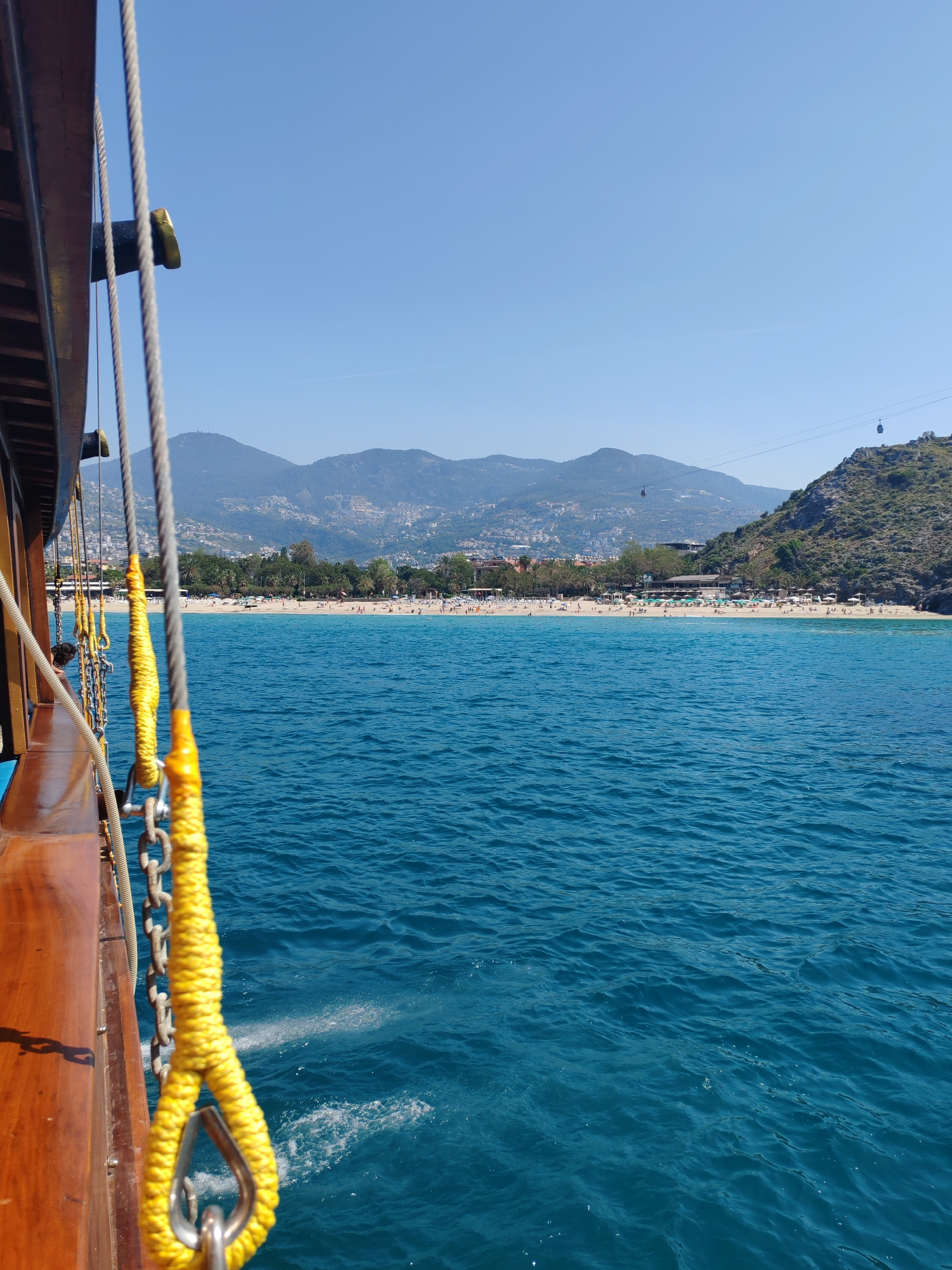 View from a wooden boat on a body of turquoise water toward a sandy beach occupied by many people, likely tourists, in Alanya, Turkey. Mountains are visible in the background. The beach is lined with palm trees and buildings, and a cable car can be seen near the top right corner. In the foreground, part of the boat is visible, including a wooden plank and yellow ropes connected to metal chains. The sky is clear and blue.