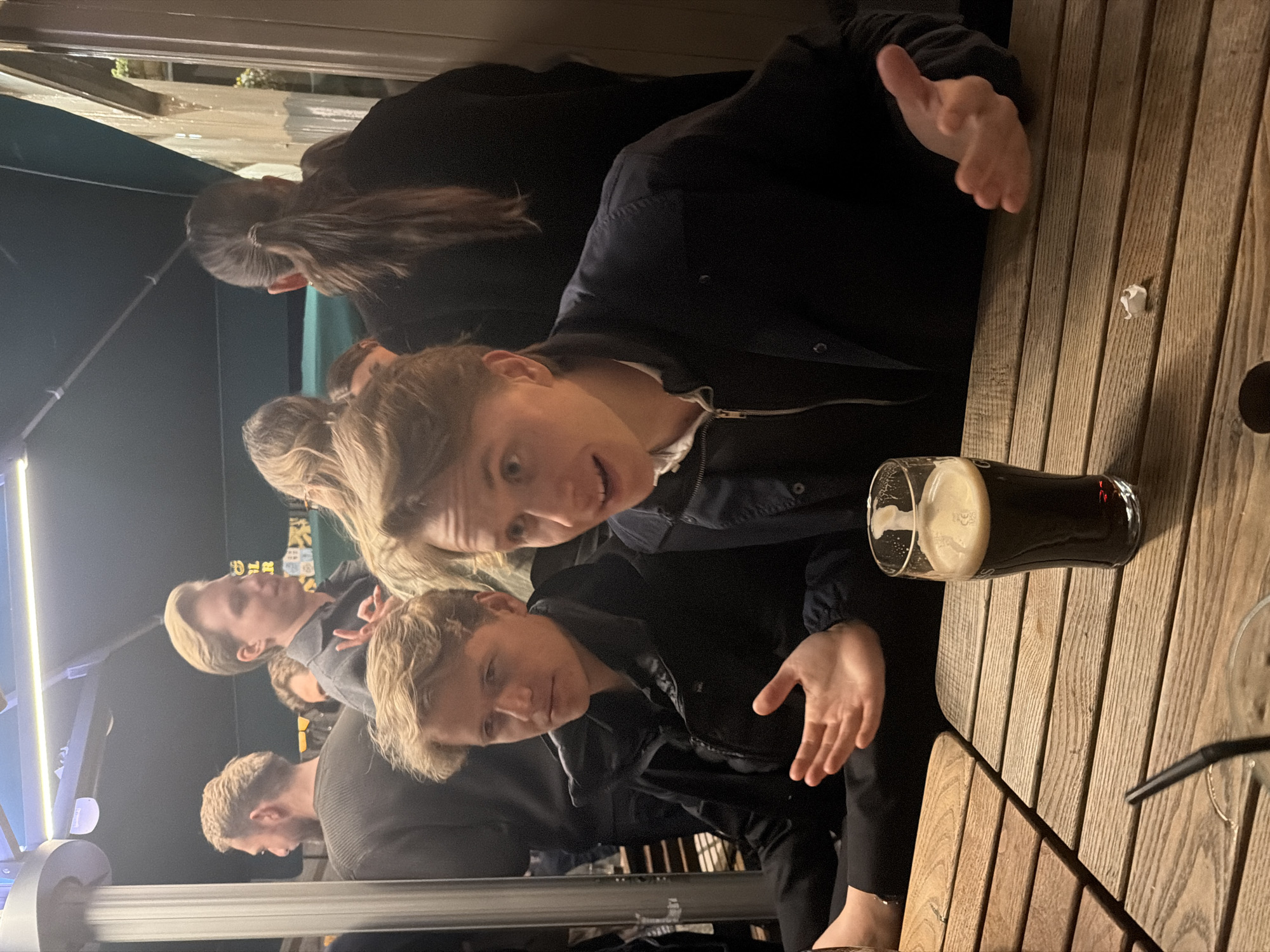 A group of people are sitting at a wooden table outdoors, likely at a pub or bar. At least one person is holding a pint of dark beer. The location is likely London, United Kingdom.