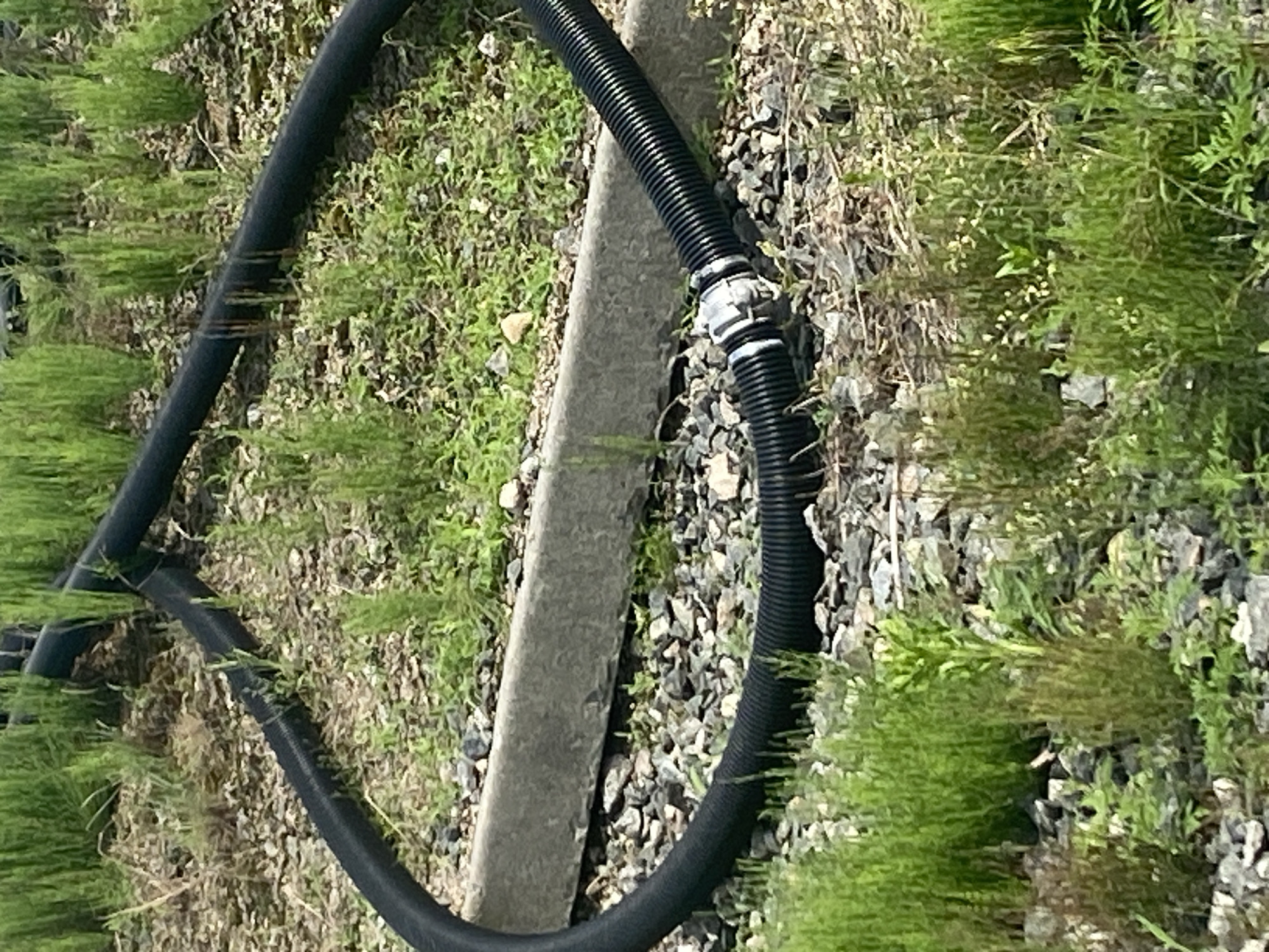 The image depicts a black flexible corrugated pipe, possibly a drainage hose, lying on the ground. The pipe is forming a loop, with a silver coupling connecting two sections. The ground is covered with gravel and scattered green vegetation, suggesting an outdoor environment. A concrete barrier runs horizontally across the middle ground. The scene appears to be located outdoors, possibly in a rural or undeveloped area, with no distinct urban landmarks visible to confirm a location in Rozhny, Ukraine.