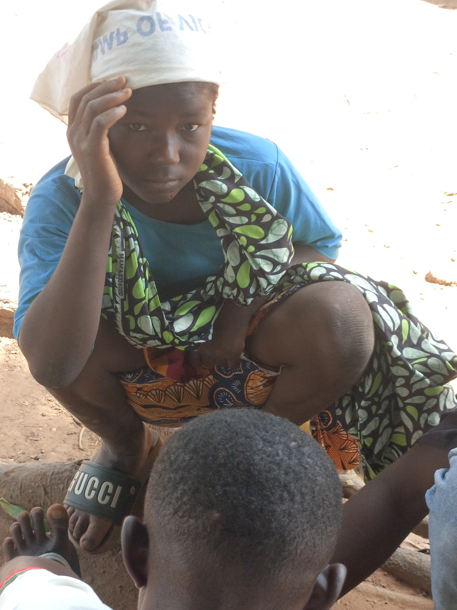 An adolescent girl in Akwanga, Nigeria, is captured in a candid pose, squatting outdoors on what appears to be a rocky or earthen surface. She is wearing a blue t-shirt, and a patterned fabric draped around her. On her head, she has a light-colored head covering with lettering visible. A "GUCCI" branded slide sandal is visible on one foot. In the foreground, the back of another person's head obscures part of the view. The overall environment is sunlit, suggesting a daytime setting in Akwanga, Nigeria.