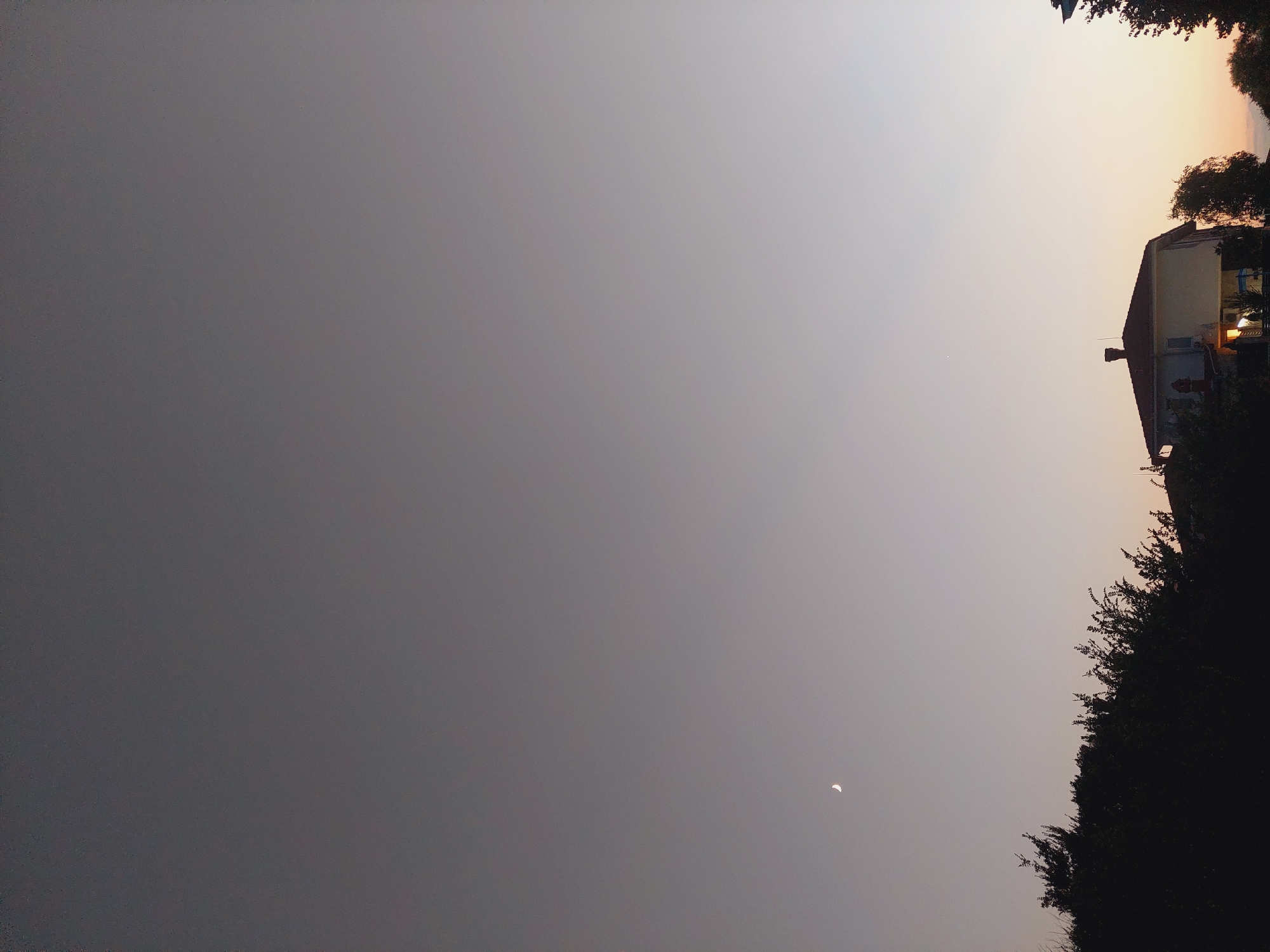 The image shows a dusk or dawn scene with a clear sky transitioning from a dark gray at the top to a lighter, warmer hue near the horizon. A crescent moon is visible in the upper left quadrant of the sky. Silhouetted against the lighter sky are the tops of dark green trees and foliage in the foreground, obscuring the lower part of the scene. A building, likely a house, with a visible roof and chimney, is situated in the lower right quadrant. Lights are illuminated within the house, casting a warm glow. To the right of the house, more dark foliage is visible, and the very edge of another building can be seen. The overall impression is a peaceful, residential view in Sozopol, Bulgaria, as twilight descends or begins.