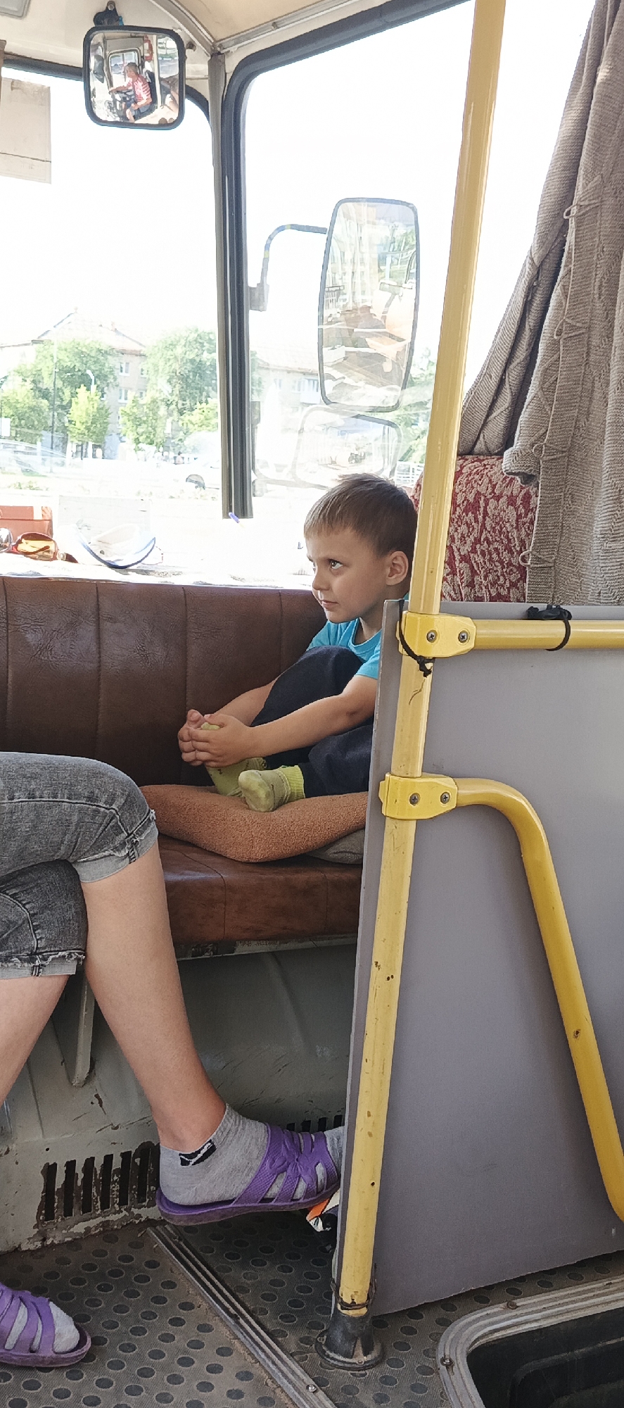 This image depicts the interior of a bus in Chaikovsky, Russia. The primary subjects are a young boy and part of another person's legs. The boy is seated on a brown, tufted bench, wearing a blue t-shirt and dark pants. He is looking out the window with a thoughtful expression, his knees drawn up and his hands resting on his legs. His feet are clad in light green socks. Adjacent to him, to the left, are the legs of another person wearing denim shorts and gray socks with purple sandals. The floor of the bus is a textured gray material.
Prominent objects include yellow metal poles and railings, part of the bus's interior structure. A patterned curtain is visible on the right side of the image. In the background, seen through the windows, is an outdoor scene with buildings and trees, suggesting a daytime setting. A small rearview mirror mounted on the windshield reflects a person driving the bus, wearing a striped shirt.
The overall impression is one of a quiet moment on a bus journey. The sunlight streaming in suggests it is daytime, possibly a warm day given the clothing. The scene appears to be set in a residential or suburban area based on the background view. There is no visible text in the image.