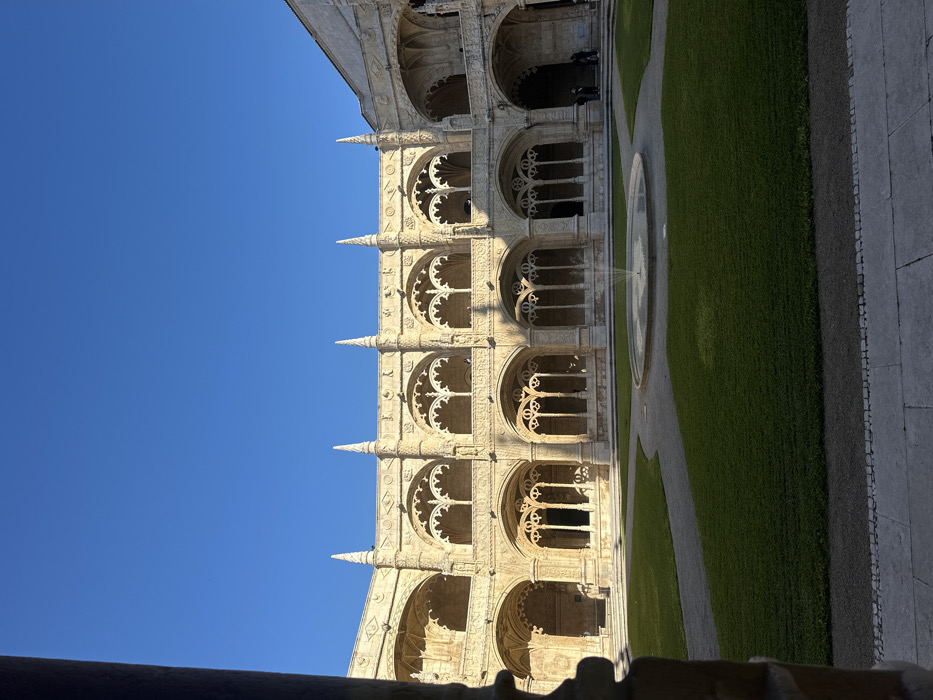 The image depicts the two-story arcaded cloister courtyard of the Jerónimos Monastery (Mosteiro dos Jerónimos) in Belém, Lisbon, Portugal, viewed from a shaded corner of the lower arcade. The architecture is characterized by elaborate light-colored limestone in the Manueline Gothic style, featuring intricate tracery, decorative balustrades, and spires topping the upper story's facade.
The central courtyard contains a manicured green lawn segmented by winding gravel paths that converge around a circular stone fountain emitting a single jet of water. Strong, low-angle sunlight illuminates the upper sections of the cloister and portions of the courtyard, casting defined shadows across the lower arcade and grass. A clear, cloudless blue sky dominates the upper half of the image.
Several individuals are visible within the shaded lower arcade on the far right side of the courtyard, appearing to observe the surroundings. One additional figure may be discernible on the upper right level. No distinct actions or interactions are clearly observable. The immediate foreground shows a shadowed stone column fragment from the viewer's perspective.