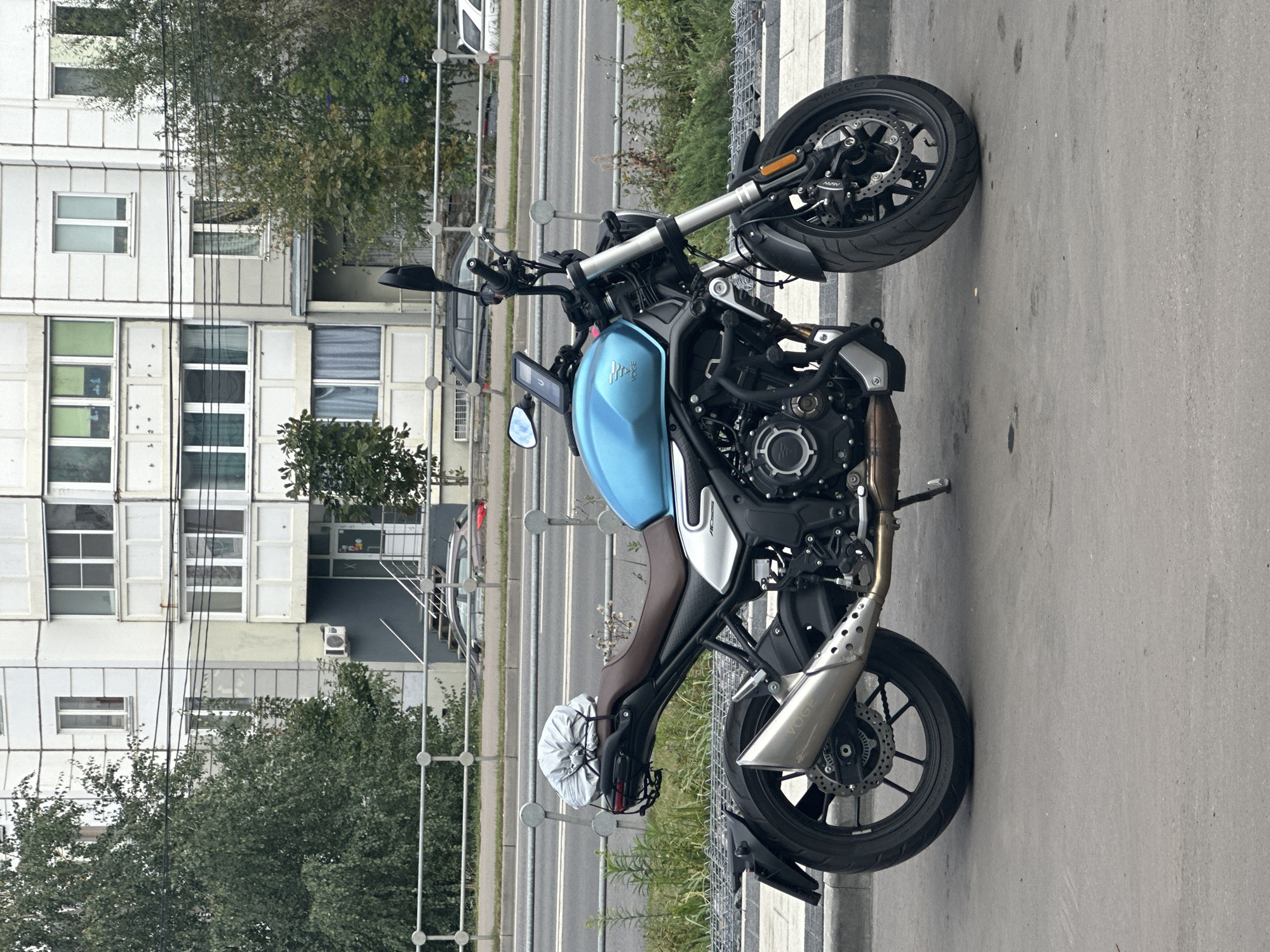 The image depicts a blue Voge motorcycle parked on a paved surface, likely a street or sidewalk, in front of a multi-story apartment building. The motorcycle is positioned in the right half of the frame, showing its front and side profile. It has a brown seat, a silver exhaust pipe, and a small silver-covered object on its rear rack. In the background, the apartment building features a light-colored facade with numerous windows and balconies. A metal fence runs horizontally across the middle ground, separating the motorcycle from a road with a few cars visible. Green trees and shrubs are present on either side of the road. The overall scene appears to be a typical urban environment. Based on the provided information, this scene is located in Podolsk, Russia.