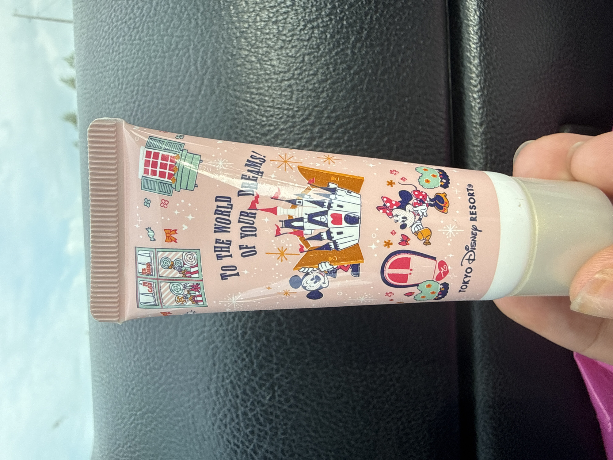 A close-up shot shows a person's hand holding a pink tube of hand cream. The tube is decorated with cartoon images of Mickey and Minnie Mouse, a castle, and windows. The text on the tube reads "TO THE WORLD OF YOUR DREAMS! TOKYO Disney RESORT®". The background appears to be the interior of a vehicle with a dark, textured dashboard. The lighting suggests it is daytime. There is no indication of the location in Chkalovka, Armenia, in the image or its details.