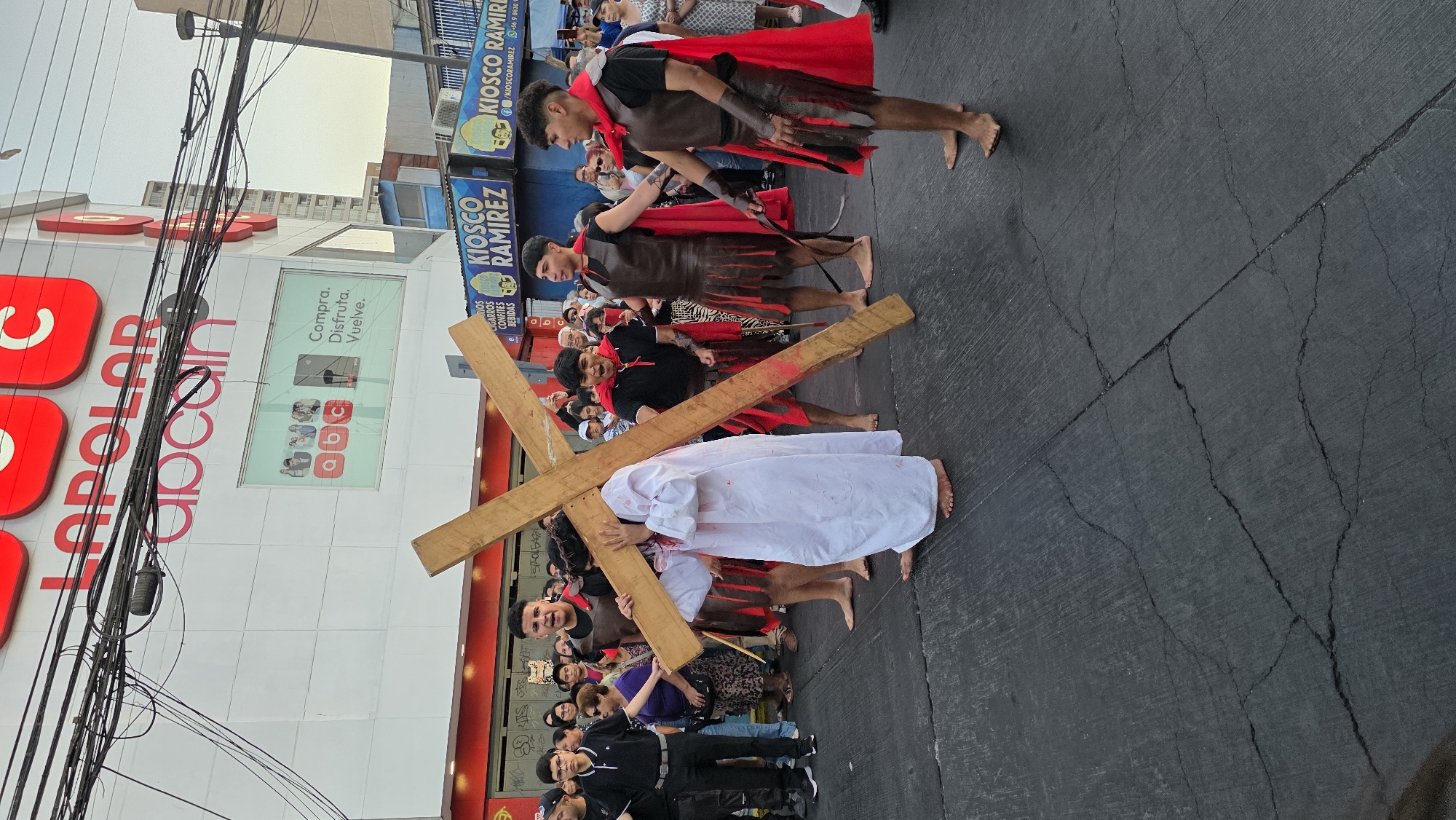Captured in an urban environment, possibly the city of Unknown, Unknown, the scene depicts a group of people participating in a religious procession, possibly a reenactment of the Stations of the Cross. One individual, dressed in a white robe, is carrying a wooden cross, signifying Jesus Christ bearing the cross. Several individuals in Roman soldier costumes, complete with red capes, are seemingly supervising or guiding the procession. A crowd of onlookers is present, observing the event. The procession is occurring on an asphalt street, with buildings and overhead power lines visible in the background. Buildings, including a large store front with red and white signage reading LOPOLAR and a window display featuring the letters abc and a sign advertising KIOSCO RAMIREZ, suggest a commercial area. The setting indicates a public space within an urban context.
