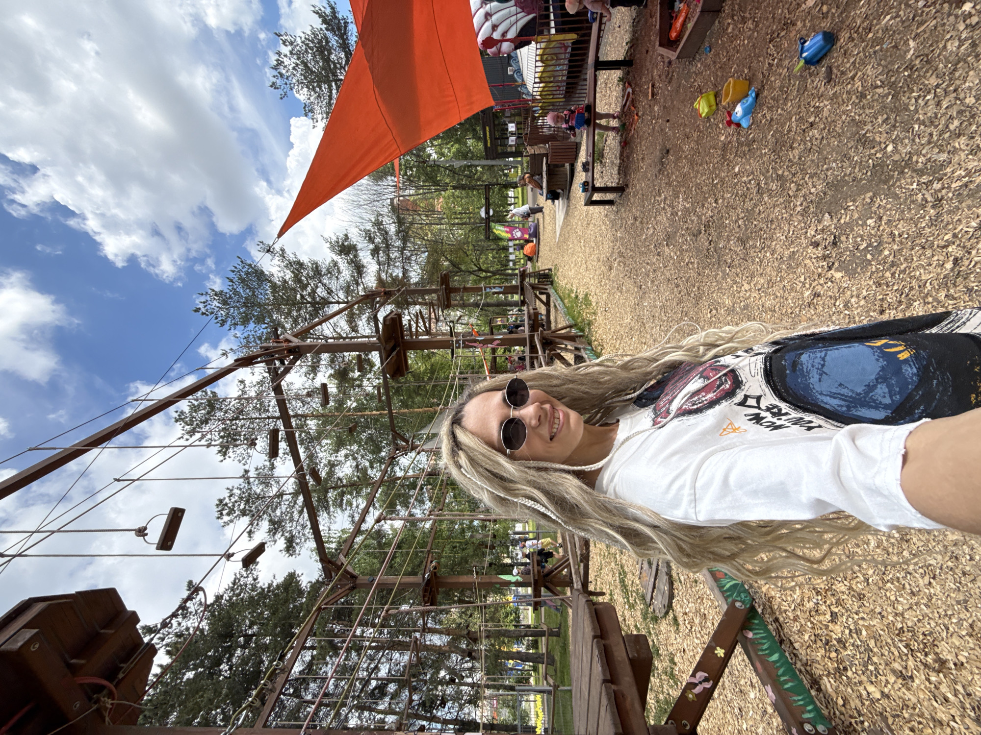 The image captures a smiling woman in a selfie-style shot at an outdoor adventure park on a partly cloudy day. The woman, positioned on the right side of the frame, has long, wavy blonde and light brown hair with braided sections. She wears dark, circular sunglasses and a white t-shirt featuring a colorful, abstract graphic design on the front, with some partially visible, indistinct text.
To her left, a large wooden ropes course structure, complete with platforms, ropes, and ladders, extends into the background, interspersed with green trees. An orange shade sail is prominently visible in the upper middle section of the image.
The ground throughout the scene is covered in light brown wood chips. In the background, beneath the shade sail, there is a recreational area with wooden benches and scattered children's toys, including blue and yellow buckets and a blue shovel. Several other people, including children and adults, are faintly visible in the background, appearing to be enjoying the park facilities. The bright blue sky with scattered white clouds suggests a daytime setting, contributing to a cheerful and recreational atmosphere.
Location context: Unknown, Unknown