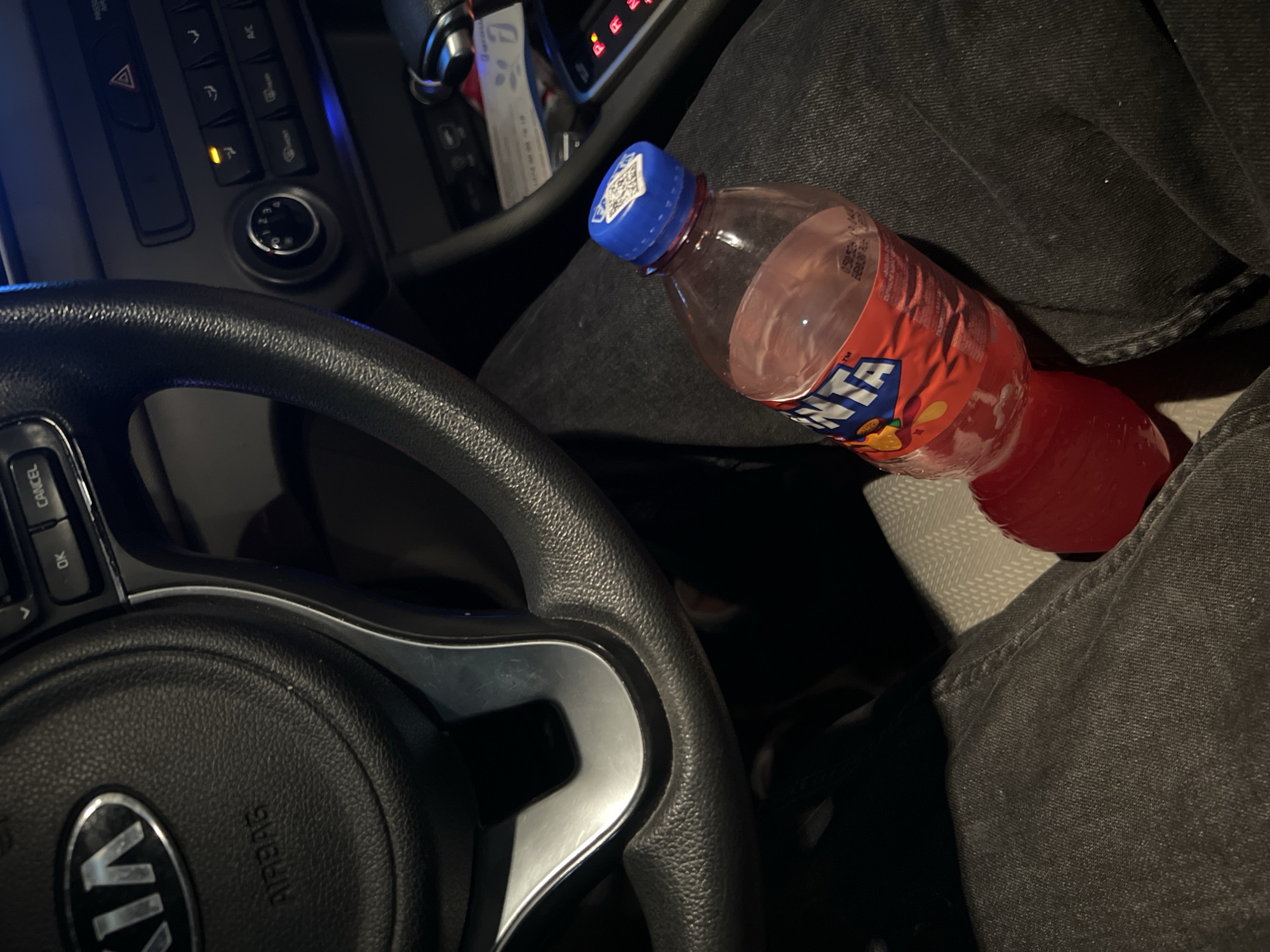 The image shows the interior of a car, specifically the driver's side. A black Kia steering wheel is prominently featured, along with part of the dashboard and center console. A bottle of Fanta soda with a blue cap rests on the lap of a person wearing dark-colored jeans. The location is not discernible from the image.