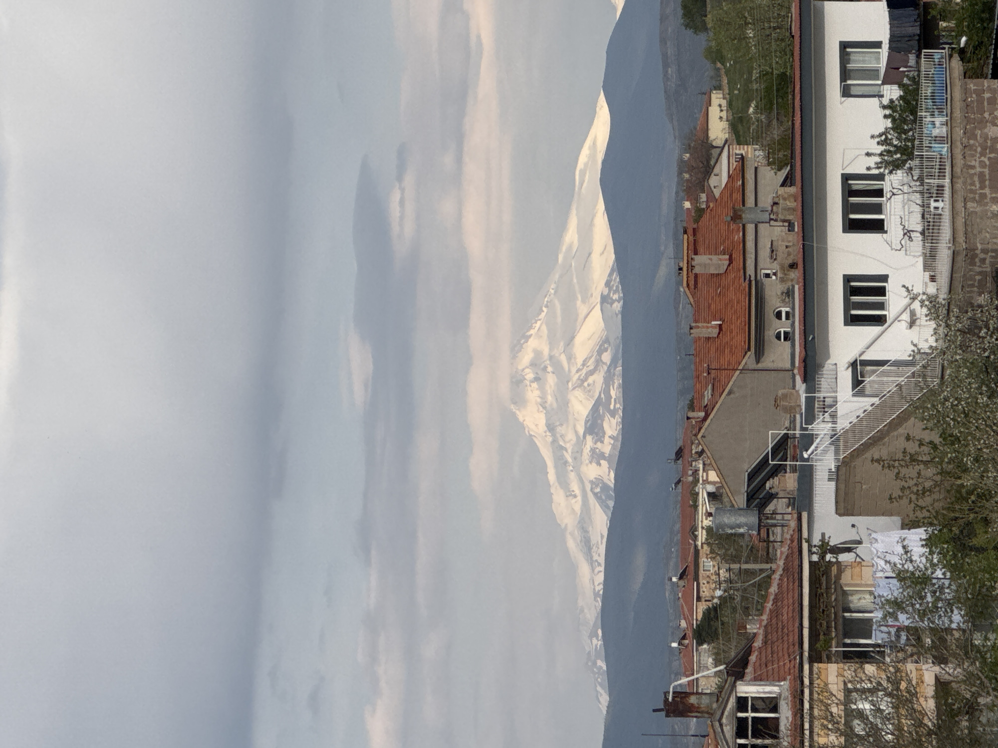 The image captures a view from Ürgüp, Turkey, showcasing a blend of urban development and natural landscapes. A snow-capped mountain serves as a backdrop to the town. To the right, a multi-story white building with dark-framed windows and a white balcony stands tall, adjacent to rooftops covered with red tiles. The building appears inhabited. The surrounding environment also reveals other buildings, varying in color and design. The sky above is partially cloudy, adding depth to the scenery.