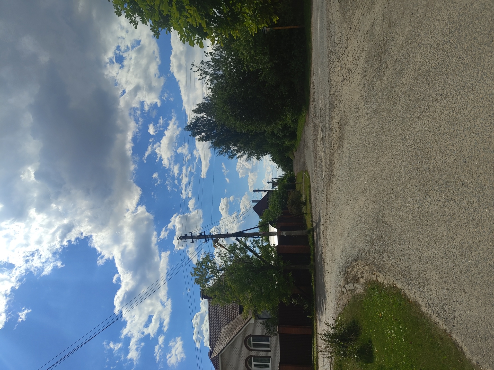 The image depicts a dirt road intersection in a rural area, likely the village of Tsibino, Russia, under a partly cloudy sky. On the left side of the road, a light-colored brick house with arched windows and a brown tiled roof is visible behind a dark brown fence. A power pole with electrical wires runs along the side of the road. To the right of the house, several trees are present. The road itself is unpaved and shows signs of wear, with tire tracks evident in some areas. Dense green foliage lines the right side of the road, and another house with a dark roof can be seen further down the road. The overall scene suggests a quiet, residential street on a sunny day.
