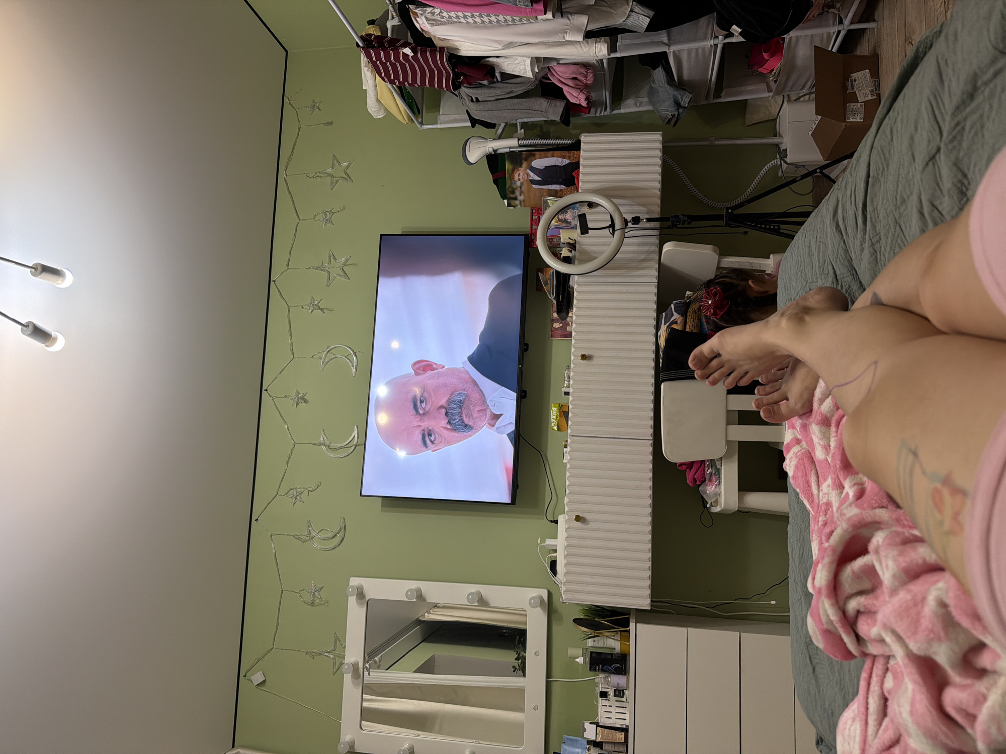 The image shows a bedroom scene. The dominant color of the walls is light green. There is a flat-screen television mounted on the wall, displaying a close-up of a man's face. Below the television is a white dresser with a ribbed front. To the left of the dresser is a white framed mirror with light bulbs around its perimeter. On the dresser are various items, including bottles and containers, and a ring light on a stand. To the right of the dresser is a clothing rack filled with clothes. In the foreground, a person's legs and feet are visible, covered partially by a pink and white striped blanket. The person appears to be lying on a bed. The overall impression is of a cozy, personal space. The location is described as Zavetny, Russia, though there are no specific visual cues in the image to confirm this location.