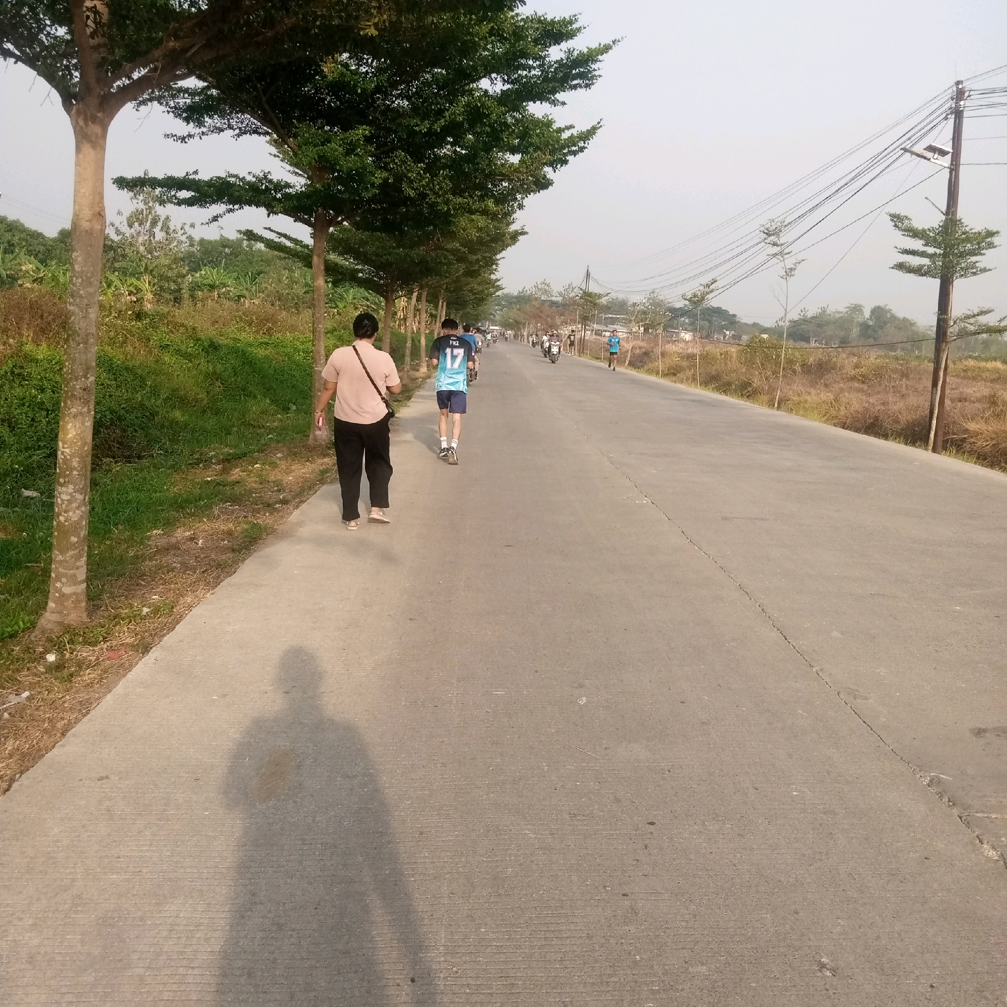 In Kendayakan, Indonesia, a paved road stretches into the distance, lined with trees and vegetation. The sky is overcast, suggesting it is early morning or late afternoon. A lone person walks on the left side of the road, facing away from the camera, wearing a light-colored shirt and dark pants. Further down the road, several individuals are engaged in recreational activities. One person in a blue and white jersey with the number 17 runs, while others are on motorcycles or bicycles, creating a scene of community activity and outdoor exercise. The environment appears rural or suburban, with houses and power lines visible in the background. The overall impression is one of a peaceful, everyday scene in a residential area.