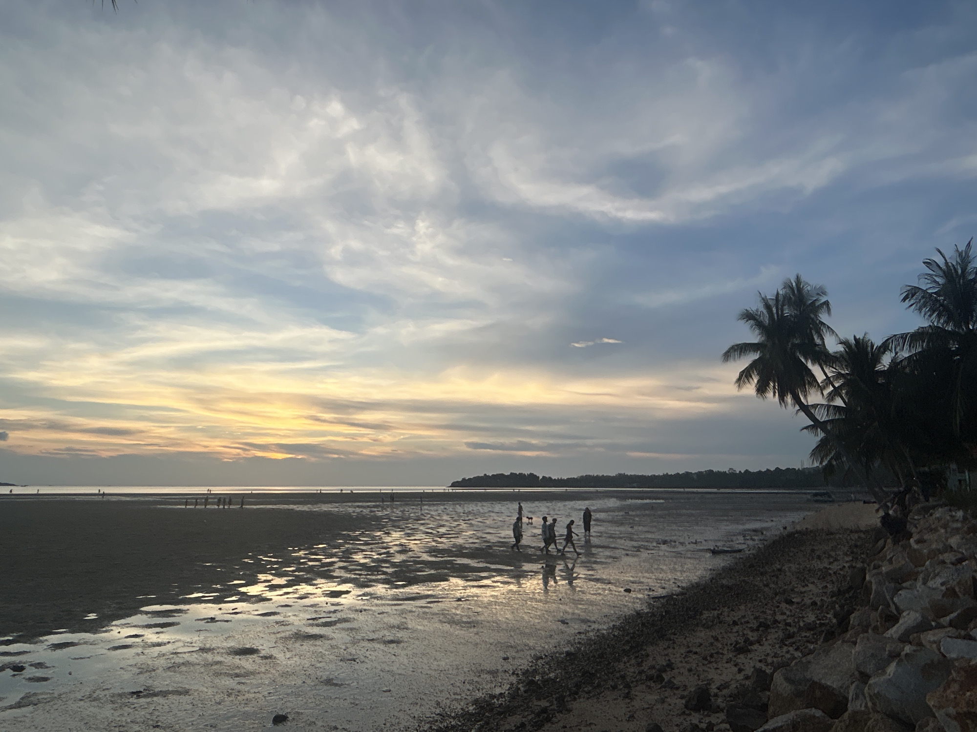 Captured at Baan Sri Thanu, Thailand, the scene depicts a low tide on a beach under a sky with hues of blue, grey, and yellow from the setting sun. A group of people is walking on the wet sand, their silhouettes reflecting in the shallow water. Palm trees are visible on the right side of the image along the shoreline. Rocks form a border on the right, near the camera. The beach extends towards the horizon where land is visible in the distance. Several people can be seen in the far left distance walking on the beach.