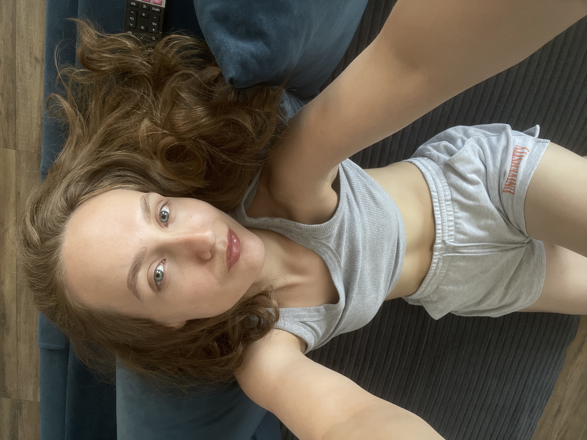 A woman is lying on a couch with a blue cushion, wearing a grey tank top and grey shorts that say "University". Her hair is brown and wavy. The location is indeterminate; there is no indication that the image was taken in Подшивалово, Russia.