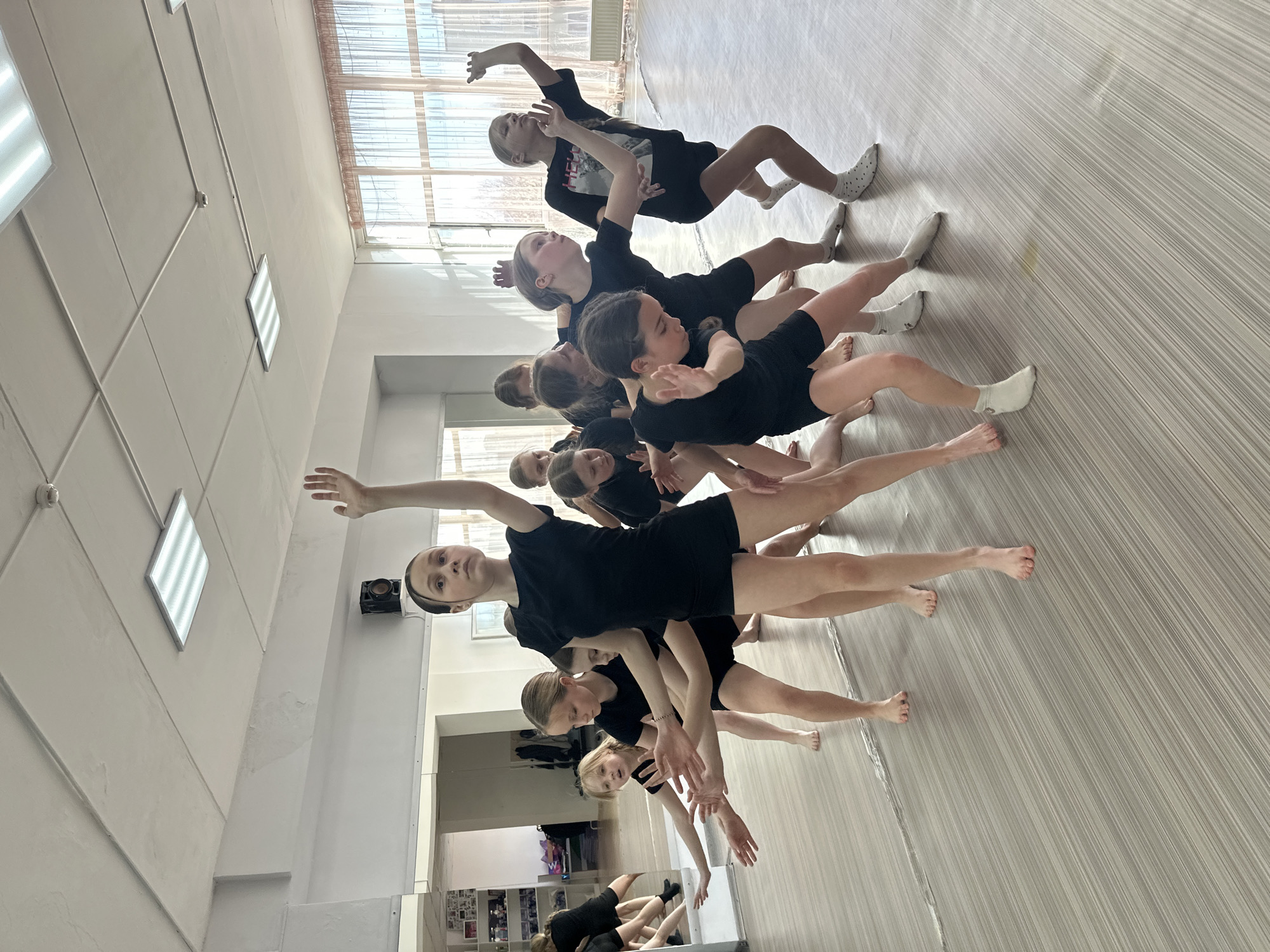 A group of approximately 11 young people, mostly female, dressed in black shorts and tops, are engaged in a dance routine inside a studio. The studio has a wall of mirrors and a large window. The floor is a light-colored linoleum with a striped pattern. The location is likely a dance studio in Votkinsk, Russia, given the assumed information. The dancers are arranged in a staggered line, with each dancer adopting a different pose, involving outstretched arms and bent legs. Some of the dancers are barefoot, while others are wearing socks. The overall scene depicts a dance class or rehearsal.