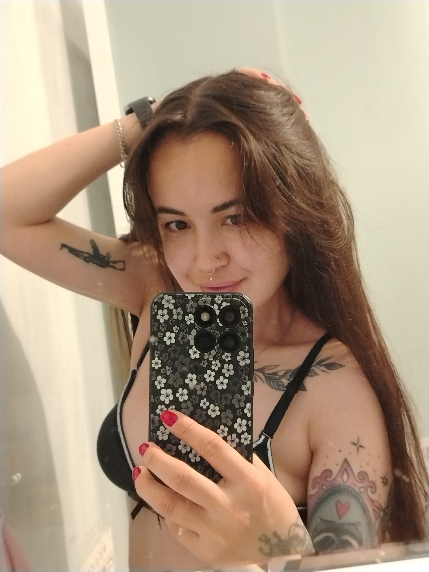 The media file shows a young woman taking a mirror selfie in what appears to be a bathroom. She has long, brown hair, a nose ring, and several tattoos on her arms and chest. She is wearing a black bra. Her right hand is holding her phone, which has a floral pattern case, while her left hand is in her hair. The reflection shows a white wall or cabinet to her left. The lighting suggests it is daytime. The woman has a neutral expression, looking directly at the camera. Visible tattoos include what appears to be a rifle on her left bicep, a leafy design across her collarbone, and a design on her right arm featuring a frame, stars, and a red heart.