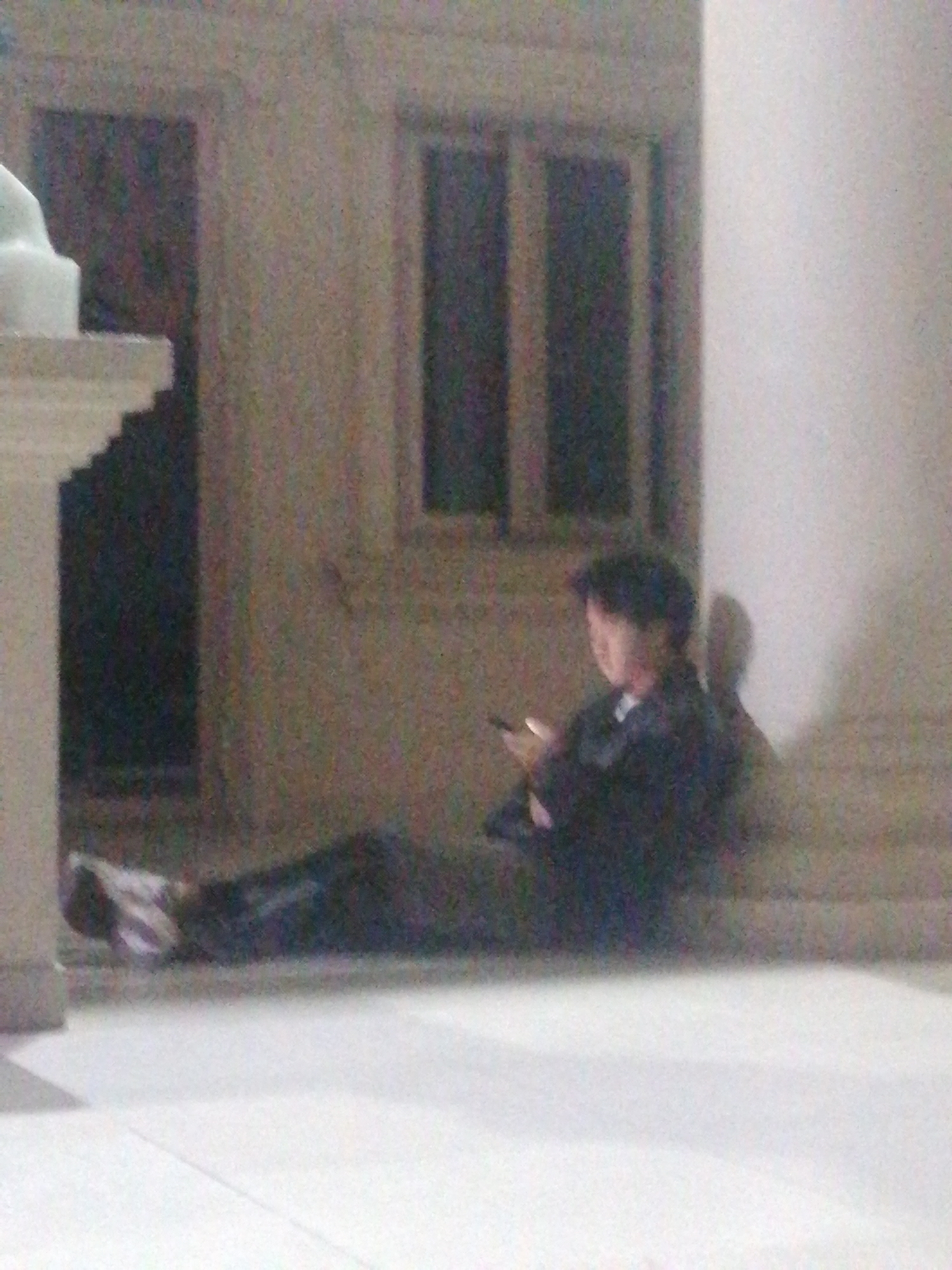 A person sits on the ground in front of a building with a light-colored pillar on the right. The person is wearing a dark jacket and has dark hair. They are holding a phone and looking down at it. The building appears to be made of concrete and has a window with vertical bars. The ground is tiled with light-colored squares. The lighting is dim, suggesting it is nighttime or indoors. The overall scene is grainy, indicating a low-quality image. The location is identified as Xuân Hương Lake in Đà Lạt, Vietnam.