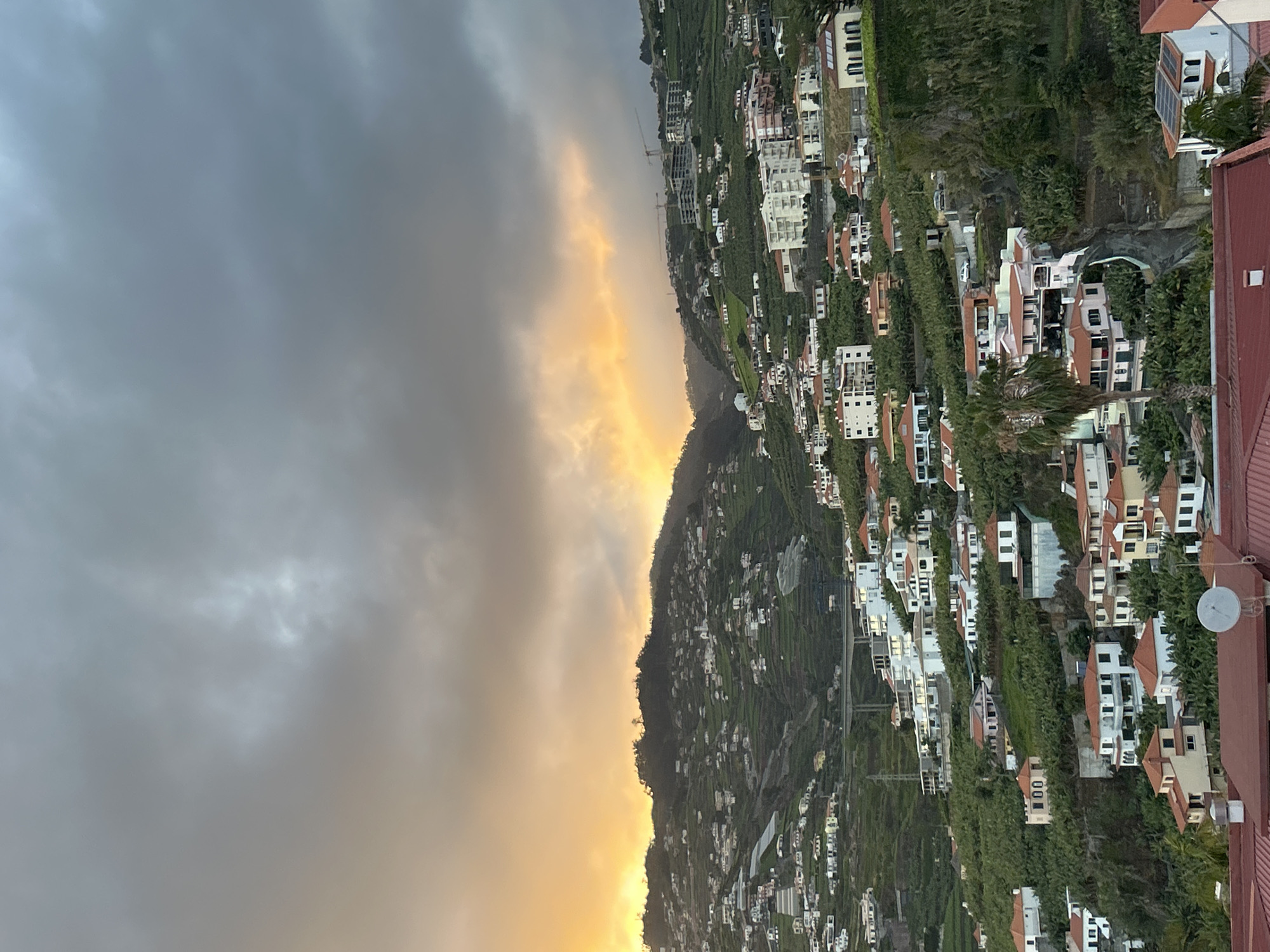 The image captures a hillside view of Câmara de Lobos, Portugal, during a dramatic sunset or sunrise. The sky is predominantly gray with a vivid orange band near the horizon. On the right side of the image, the hillside is covered with residential buildings interspersed with greenery. The houses vary in design, most featuring red tiled roofs. A satellite dish is visible on one of the rooftops in the foreground. The terraced landscape slopes steeply down towards the glowing horizon. The overall scene suggests a contrast between the urban development and the natural environment, illuminated by the atmospheric sky.
