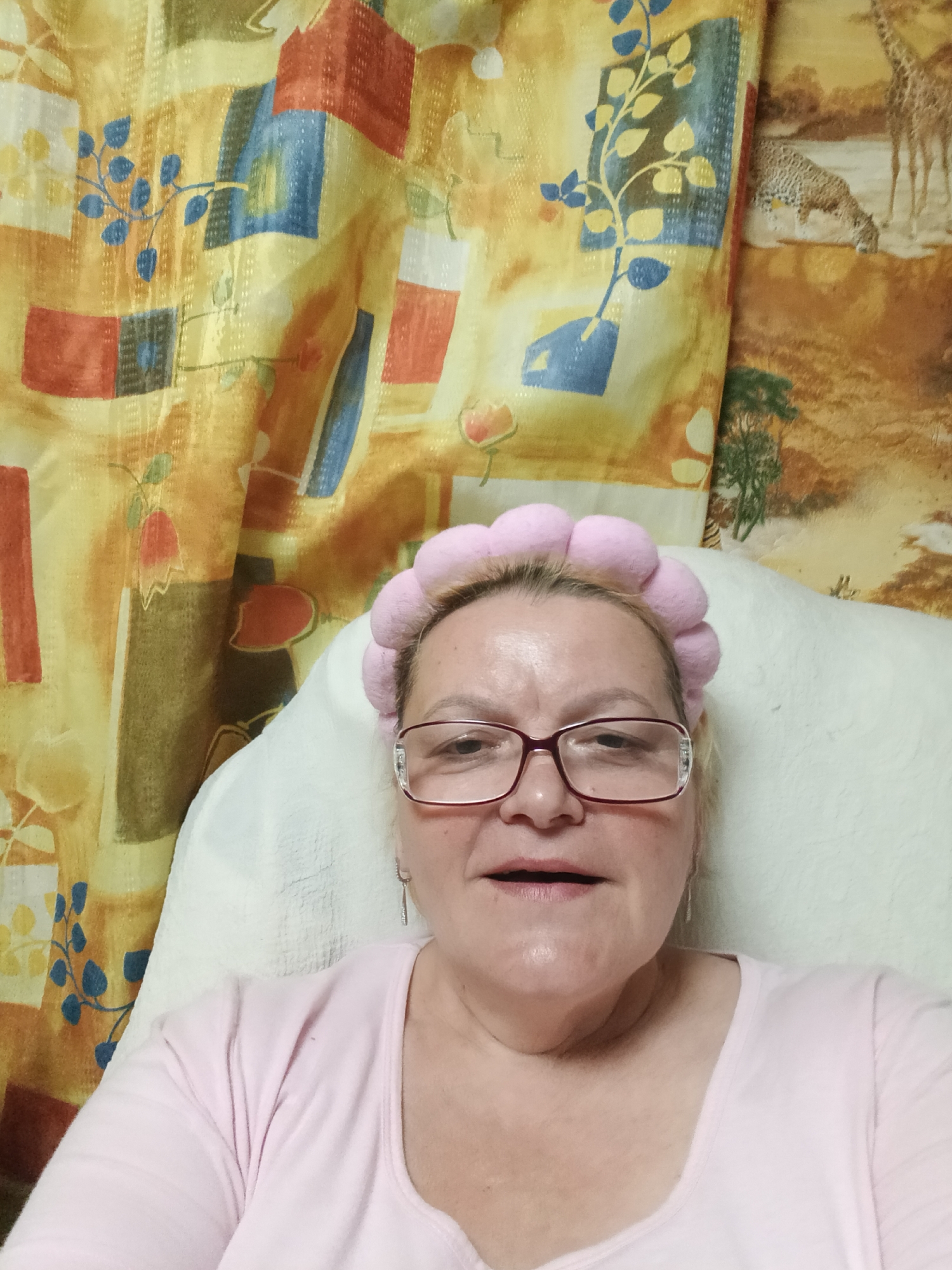 A woman wearing a pink puffy headband and glasses is seated in what appears to be a chair with a white headrest or cushion. She is wearing a light pink shirt with a scoop neck. Behind her, there is a busy patterned curtain or wall hanging in shades of yellow, orange, blue, and red. To the right of that, a portion of a wall hanging is visible with a depiction of a giraffe. There is no information in the image to indicate that it was taken in Красный Маяк, Russia.
