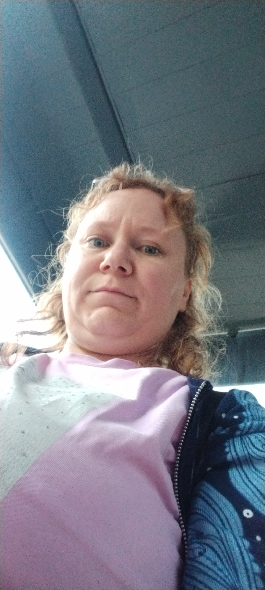 A person with reddish-blond curly hair, blue eyes, and fair skin is taking a selfie. The person is wearing a pink shirt with white and silver accents and a blue and black jacket. The background appears to be the interior of a vehicle with a dark blue ceiling. The location is unknown and cannot be confirmed as the city of Chaikovsky, Russia based on the visual information provided.