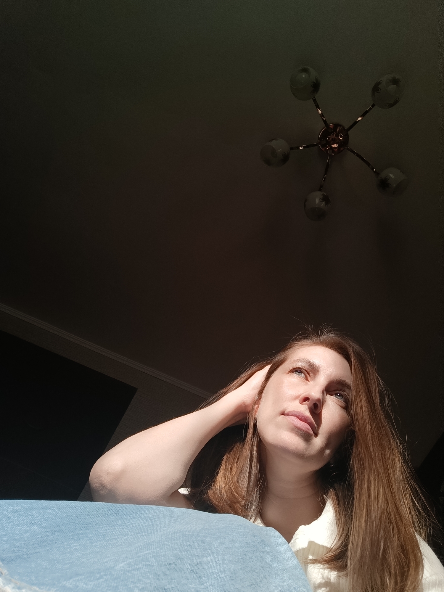 A low-angle shot indoors shows a woman gazing upward with her right hand resting on her head. She has fair skin, light hair, and is wearing a light-colored garment. Part of a blue surface is visible in the lower left. Above her, in the center of the frame, is a ceiling light fixture with six frosted glass shades. The ceiling behind is dark, with some light entering from the side. There is no evidence of the location being Фоки, Russia.